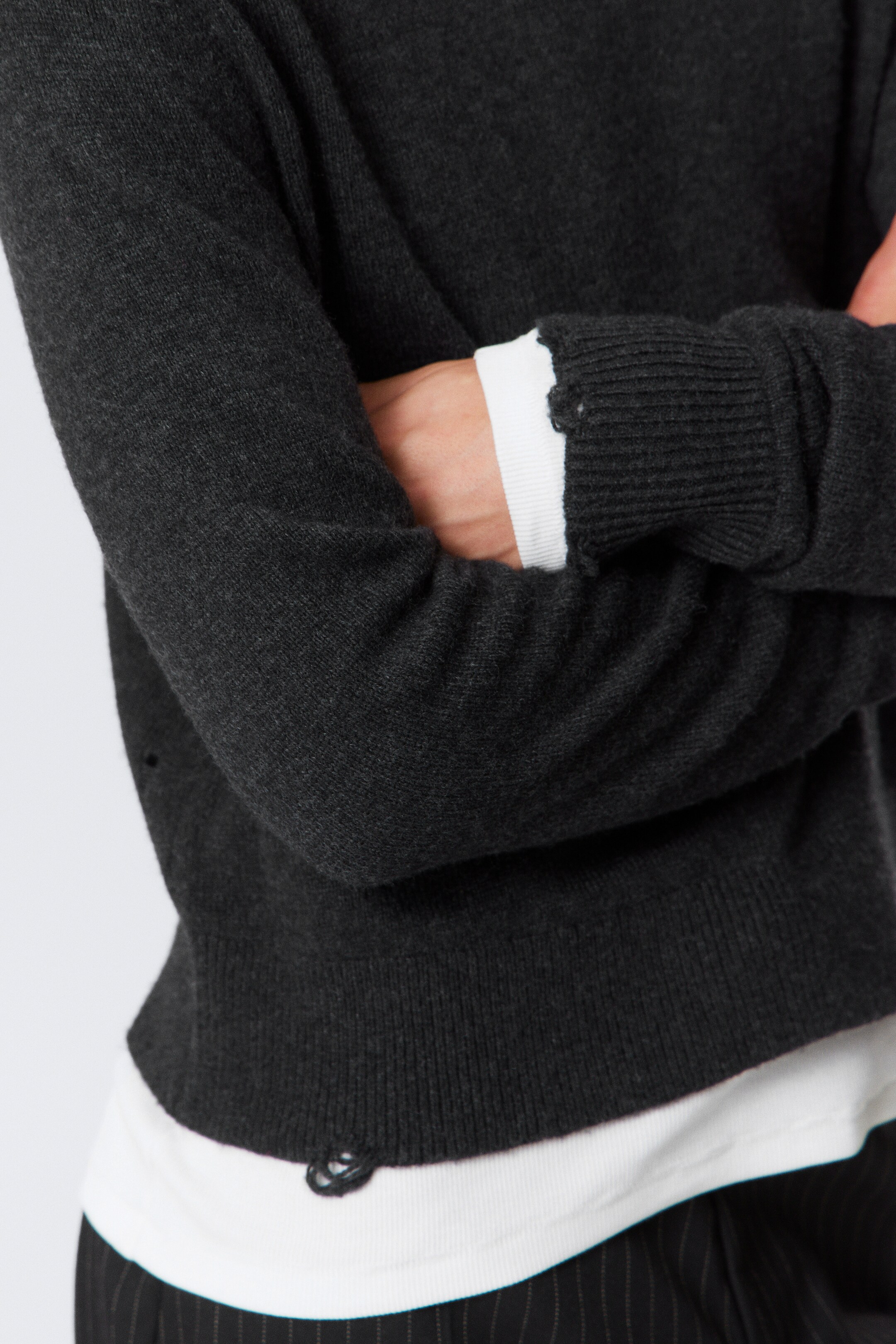 View larger image: A dark grey sweater is being presented with its arms crossed, featuring ribbed cuffs and a ribbed hem. Both the cuffs and hem are showing distressed details, with a white long-sleeved top layered underneath.