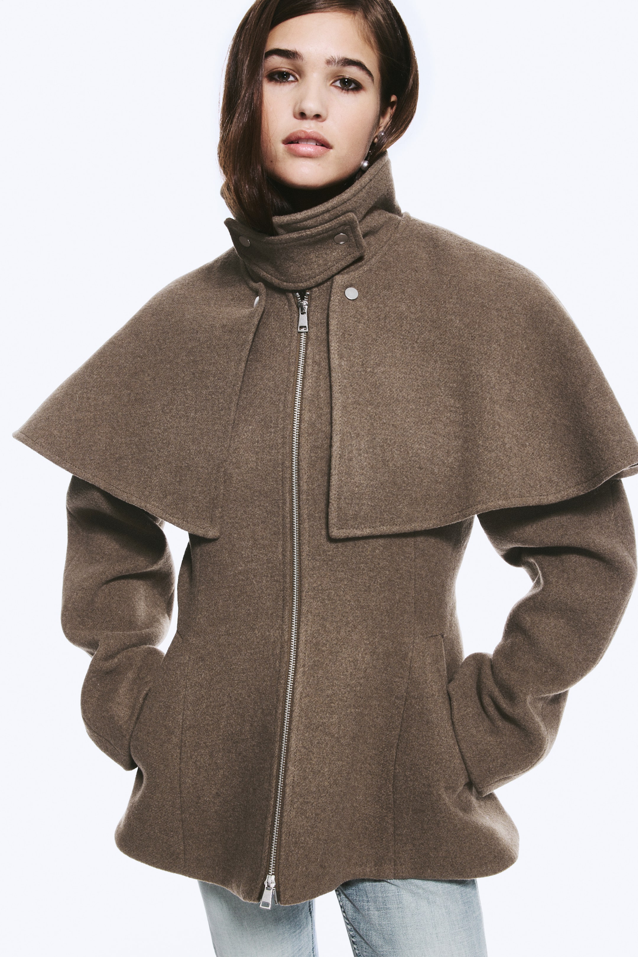 Ver imagen más grande: La modelo lleva una chaqueta de mezcla de lana marrón con cuello alto tipo chimenea, capa estructurada en los hombros y cremallera frontal plateada. La modelo está de pie con las manos apoyadas en los bolsillos laterales de la chaqueta, combinada con unos vaqueros azul claro.