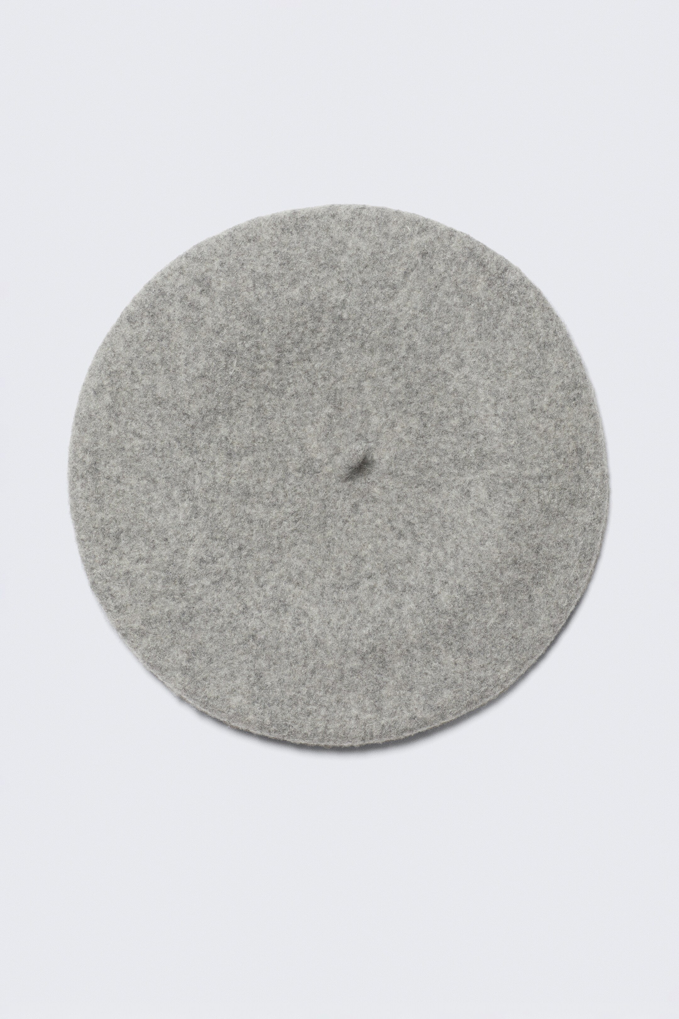 Agrandir l'image: Un béret gris clair est présenté en vue de dessus, posé à plat. Il présente une forme circulaire avec une texture douce et feutrée et un petit détail de tige en son centre.