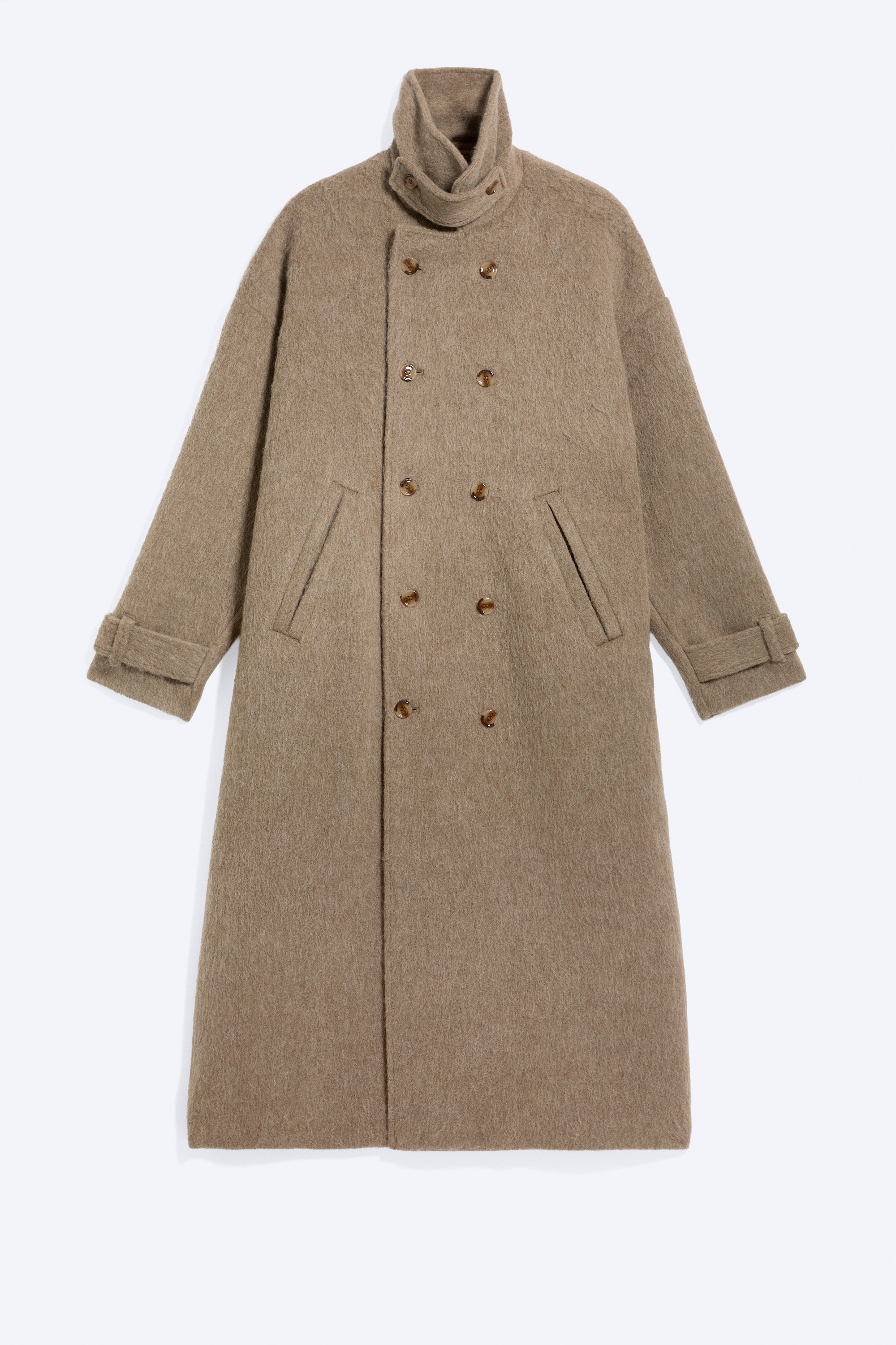 En oversized frakke med dobbeltradet lukning i en beige, hÃ¥ret uldblanding bÃ¦res af modellen. Frakken med lang silhuet har en nedfoldelig krave og knaplukning og er stylet over en mÃ¸rk hÃ¦ttetrÃ¸je og sweatpants.