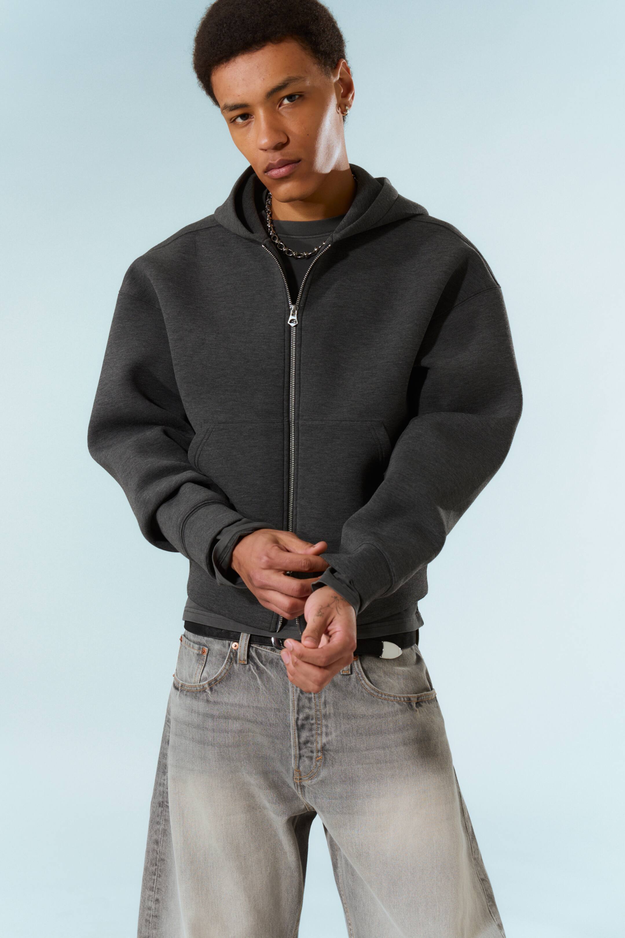 Ingrandisci l'immagine: Una felpa con cappuccio a zip intera di colore grigio scuro presenta una cerniera tono argento che corre lungo la parte anteriore, un cappuccio attaccato e una tasca a marsupio singola. Offre una vestibilità rilassata con spalle cadenti e polsini spessi, ed è stratificata sopra una maglietta scura con l'orlo inferiore visibile.