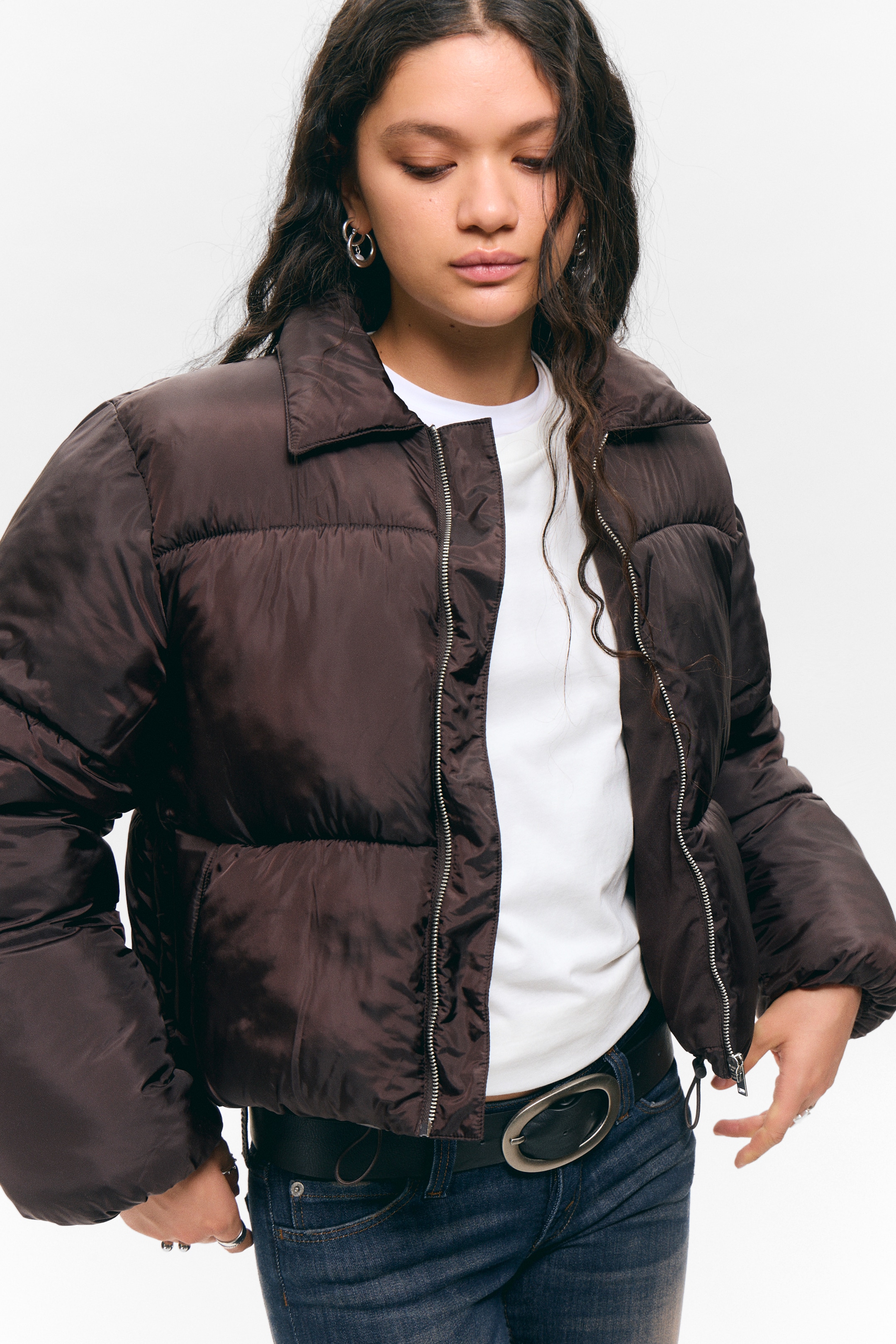 View larger image: A dark brown cropped puffer jacket is being presented from a front-facing angle, featuring a spread collar and a partially open silver zipper. The voluminous jacket is showcasing its puffed construction, with a white t-shirt visible underneath and a drawstring detail at the hem.