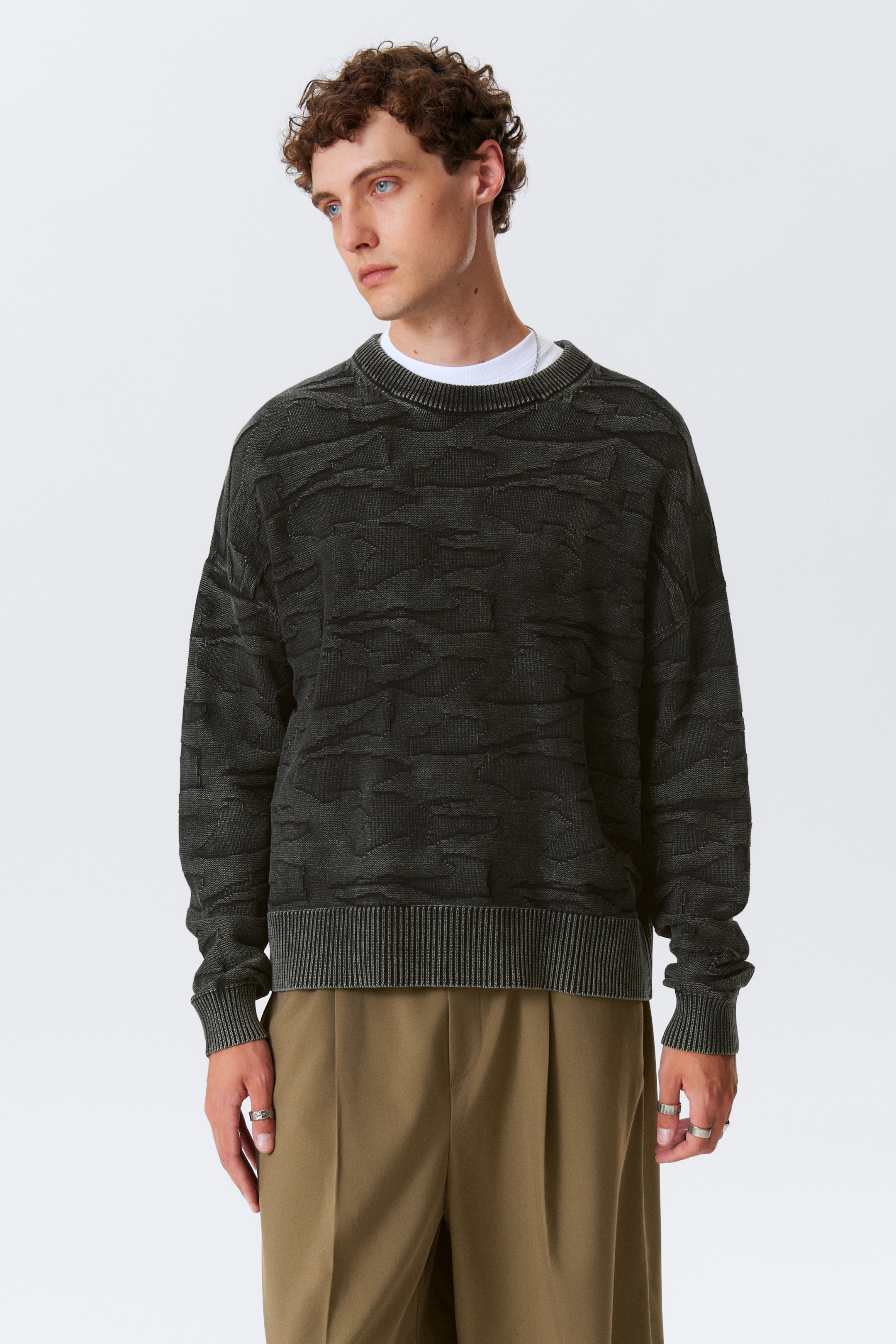 Größeres Bild anzeigen: Ein dunkelgrauer Pullover mit Rundhalsausschnitt wird aus einem frontalen Winkel gezeigt und weist ein strukturiertes, abstraktes Muster auf. Er ist mit einem gerippten Kragen, gerippten Bündchen und einem gerippten Saum gestaltet und wird über einem weißen T-Shirt mit Rundhalsausschnitt getragen.