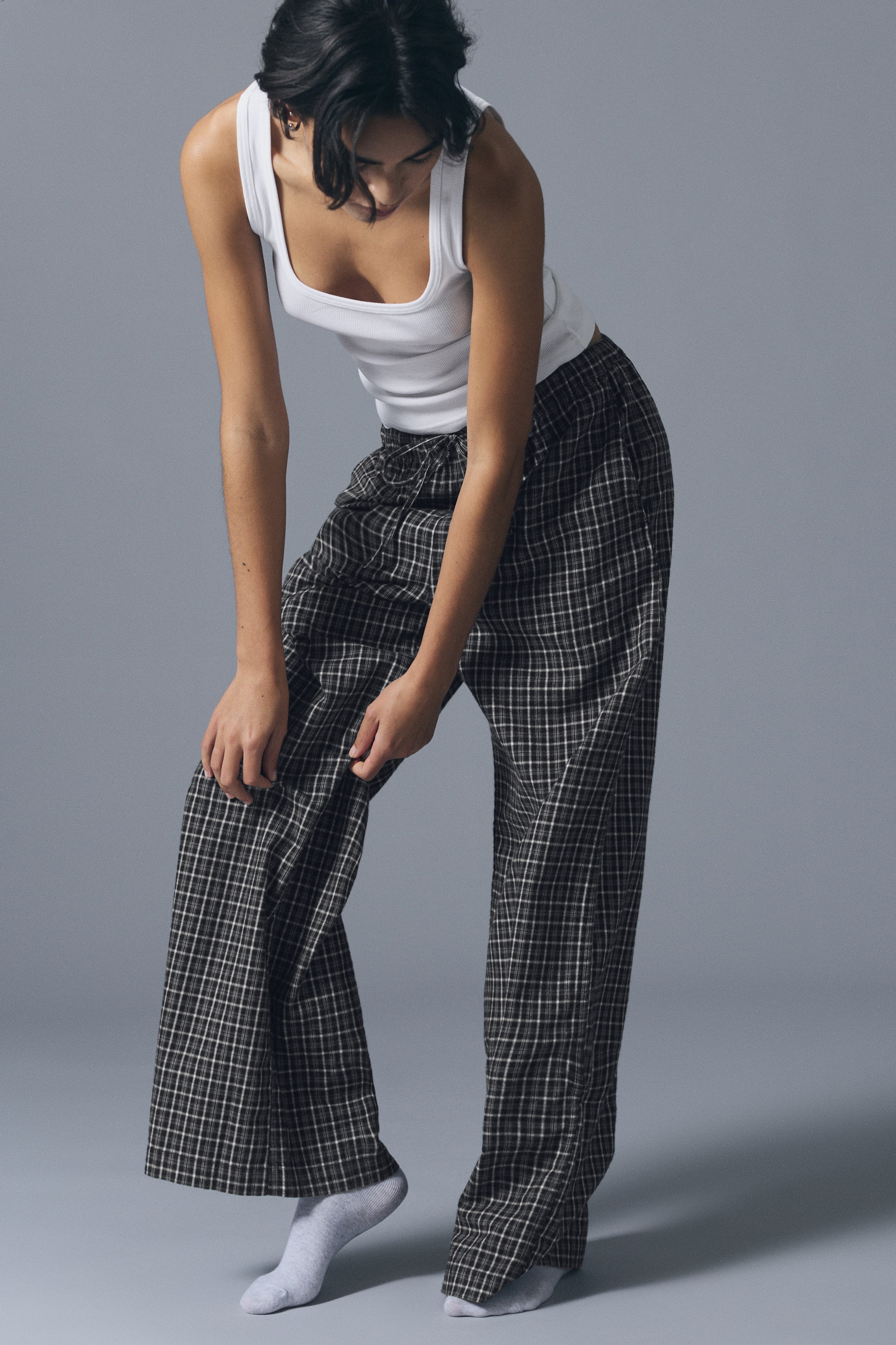 Größeres Bild ansehen: Weit geschnittene Flanell-Pyjamahose mit dunklem Karomuster, getragen von dem Model, das sich nach vorne beugt und den Stoff eines Hosenbeins hält. Die Hose mit Kordelzugbund wird mit einem weißen gerippten Tanktop und hellgrauen Socken kombiniert.