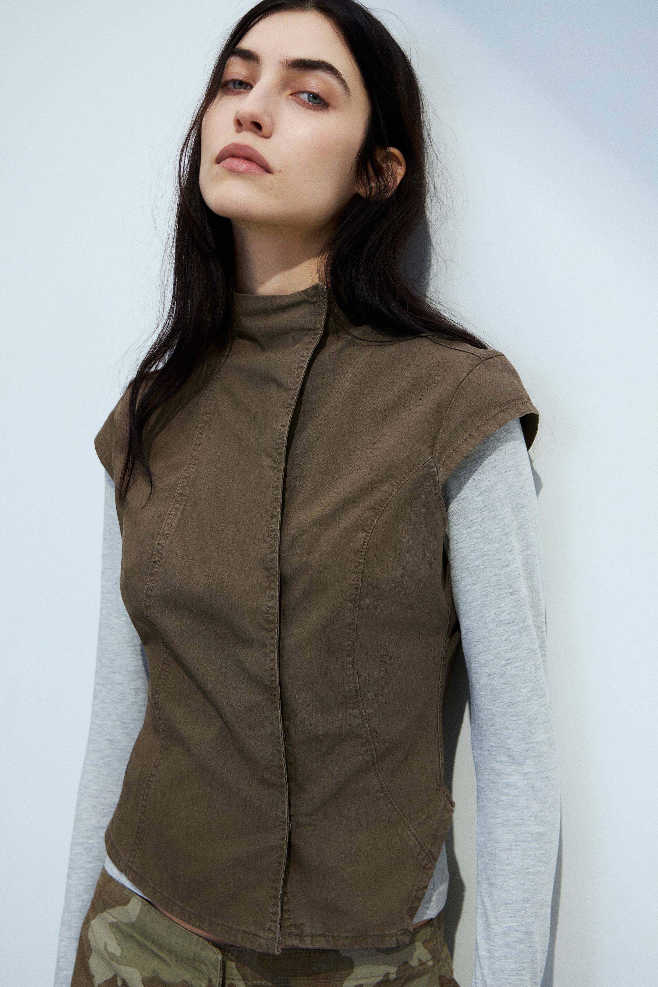 Visa större bild: En brun ärmlös väst med ståkrage, en snedställd knäppning framtill och böjda sömmar som formar dess skräddarsydda passform, bärs över en ljusgrå långärmad skjorta och presenteras i en lätt vinkel.