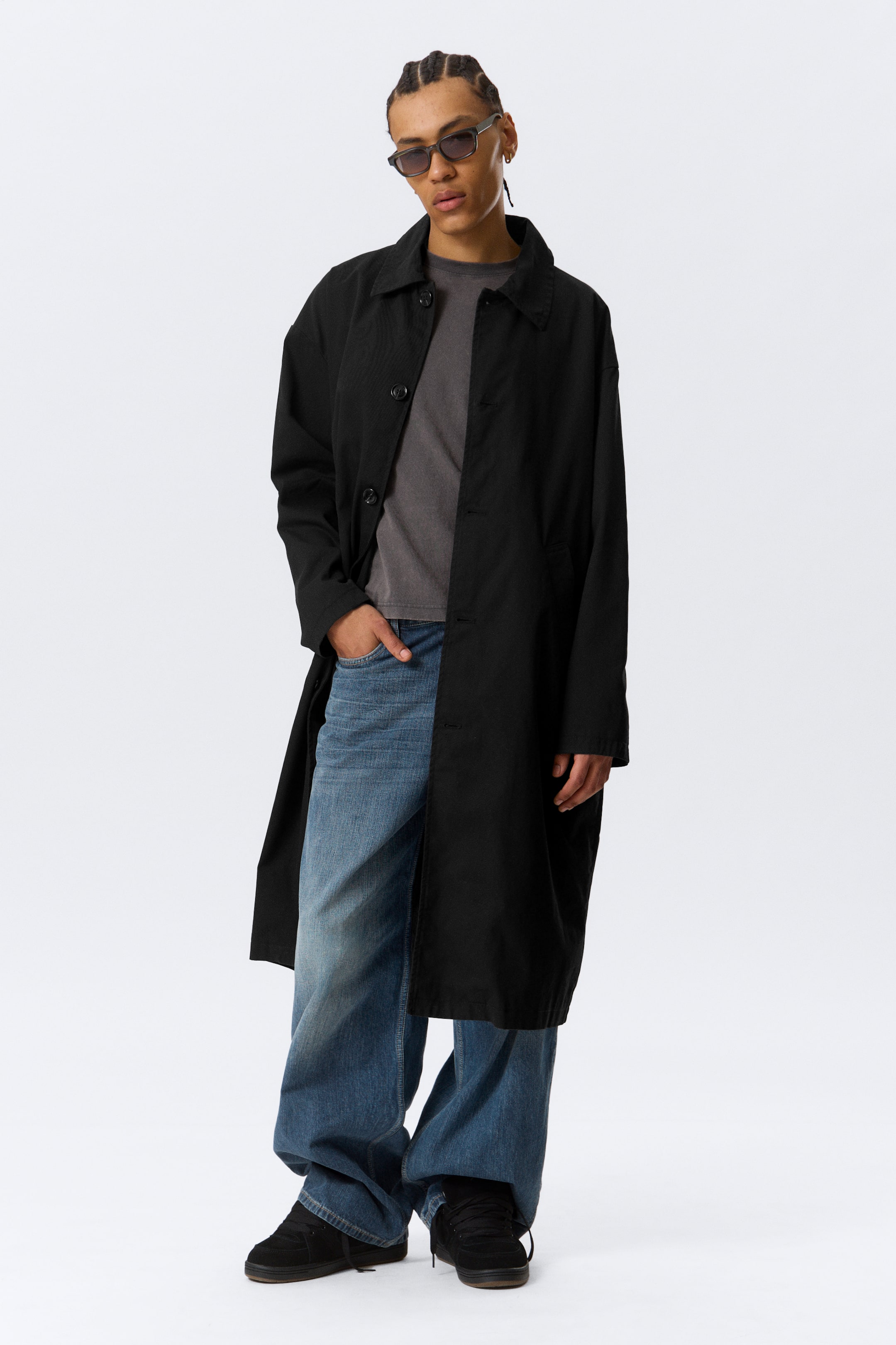 Agrandir l'image: Un manteau long noir à coupe décontractée avec un col à revers et une fermeture à boutons est porté par le mannequin. Il est stylisé ouvert sur un t-shirt gris et un jean bleu à jambes larges.