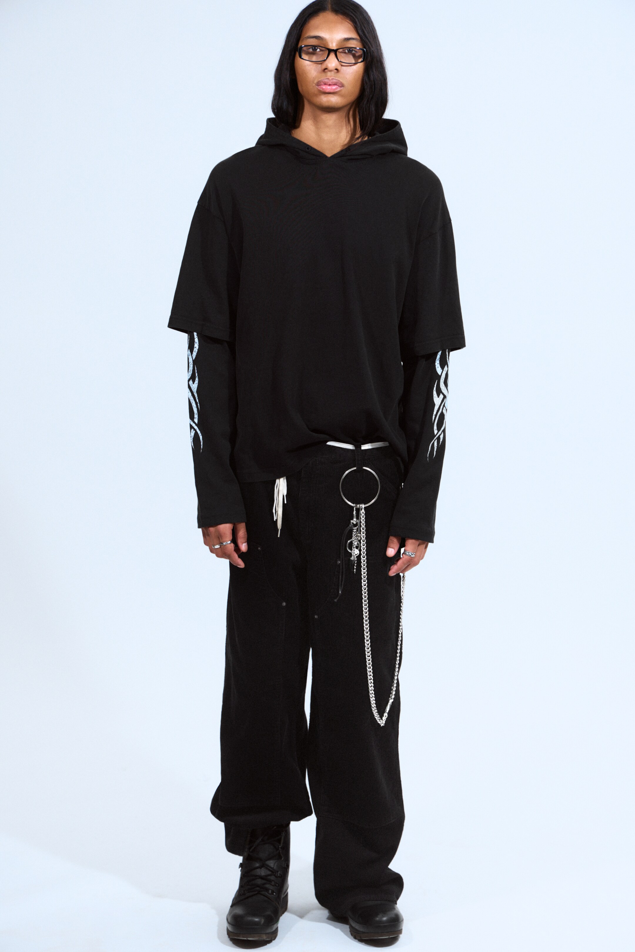 View larger image: An outfit is being presented, facing forward, featuring a black short-sleeved hooded sweatshirt layering over a black long-sleeved shirt with light blue flame graphics on its sleeves. The ensemble includes black wide-leg trousers, a thin white belt from which a silver-toned chain accessory is hanging, and black boots.