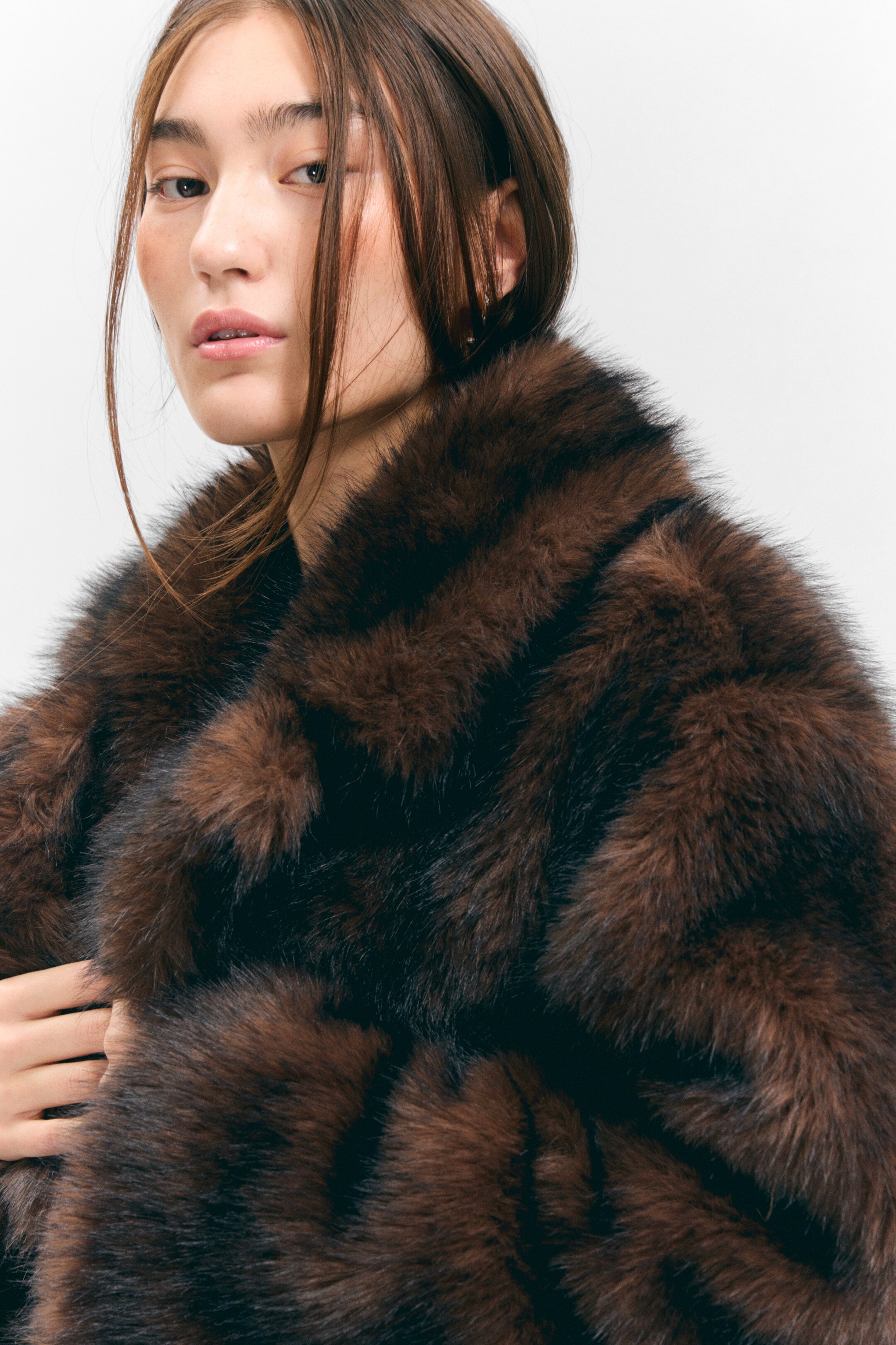 Agrandir l'image: Un manteau en fourrure brune est présenté de face, légèrement incliné, mettant en valeur sa fourrure pelucheuse aux tons variés. Le manteau présente des rayures marron foncé et plus claires sur les épaules et un large col, avec une main reposant sur la fourrure, révélant sa profondeur et sa douceur.