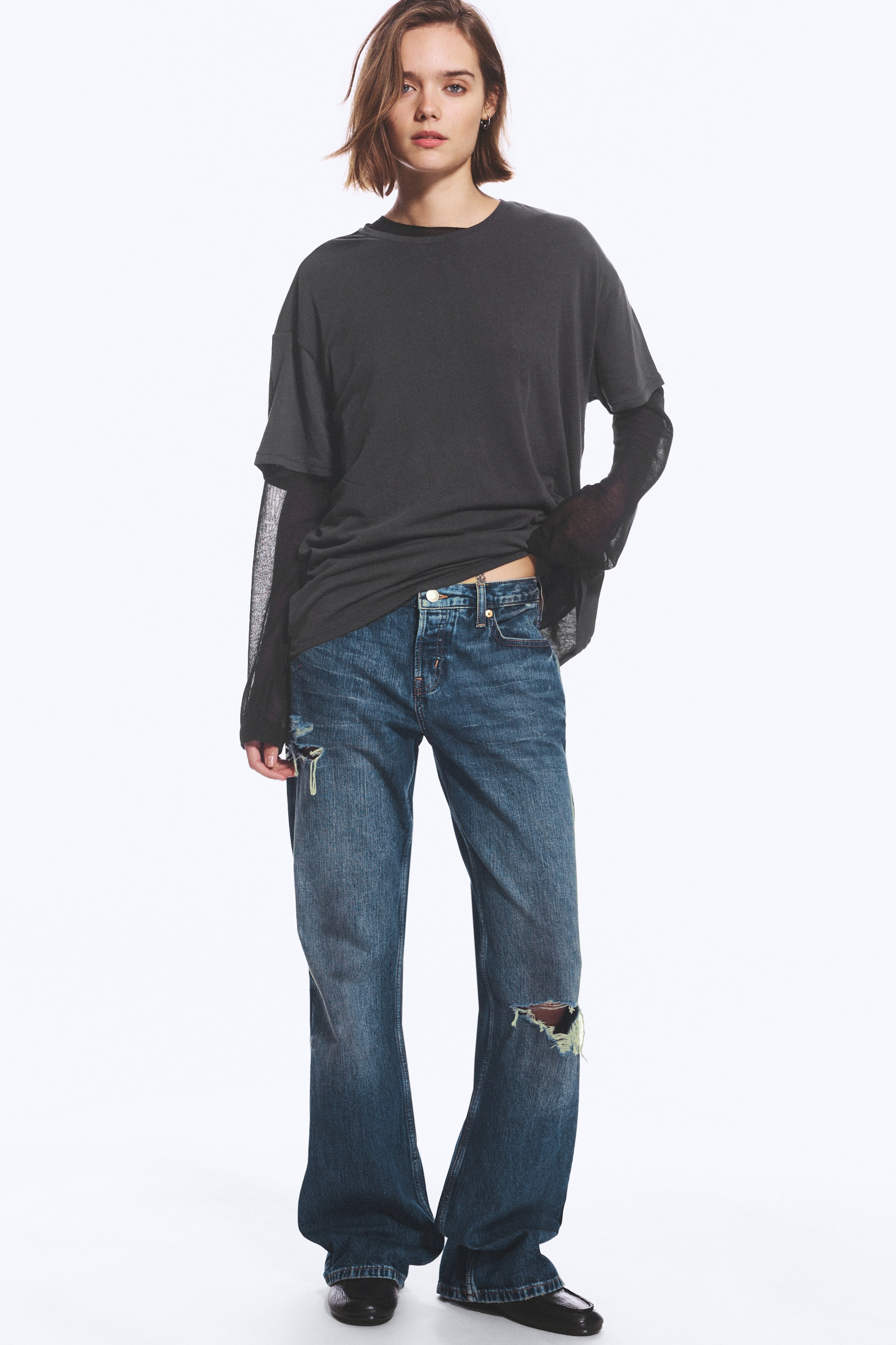 Visa större bild: Blå denimjeans med låg midja, avslappnad passform och slitna detaljer vid knäna bärs av modellen. De hellånga bootcut-jeansen är stylade med en lager-på-lager mörkgrå T-shirt och svarta skor.