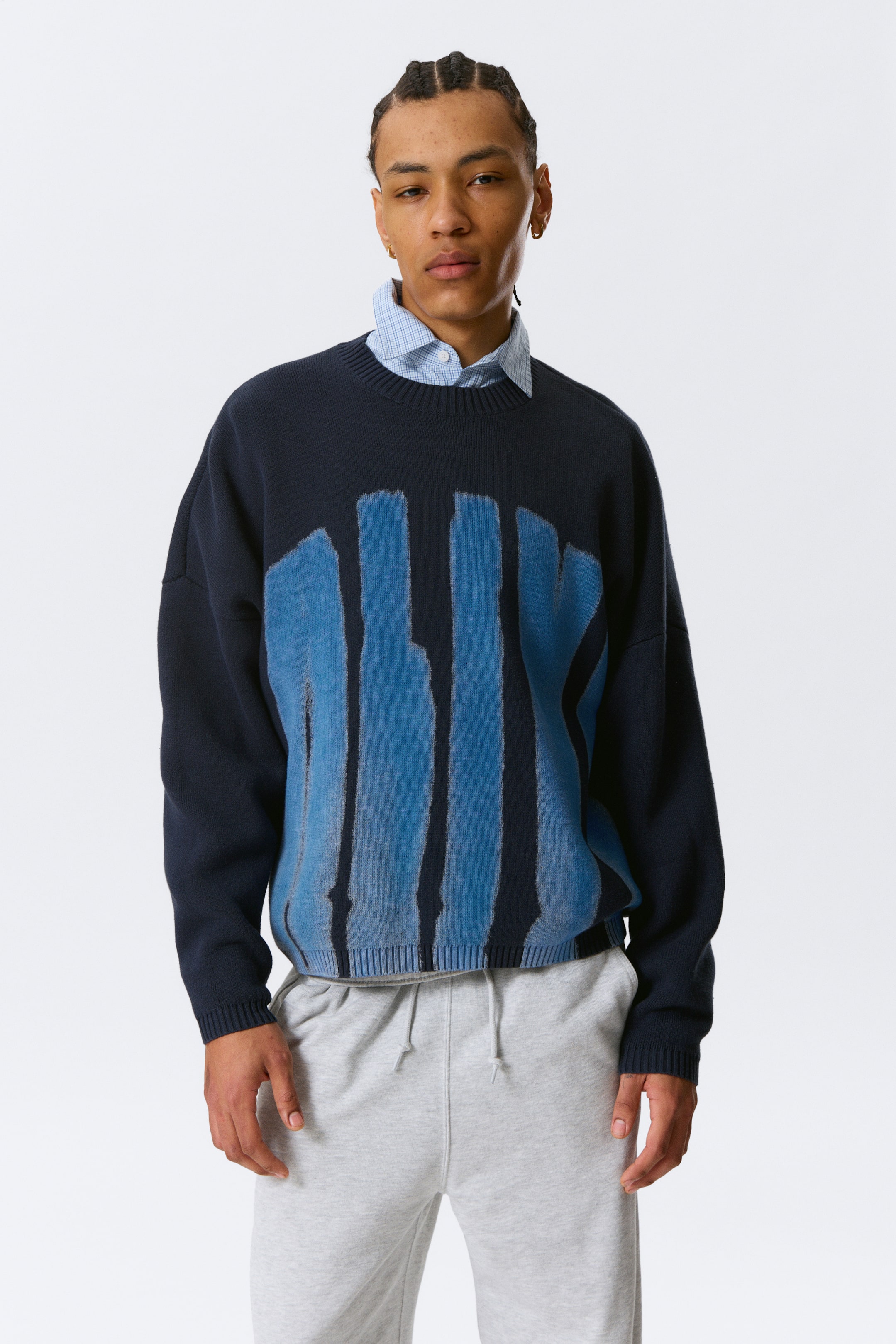 Ver imagem maior: Um camisola de gola redonda azul-marinho apresenta um design abstrato de pinceladas verticais azuis na parte frontal. Exibe punhos canelados e uma bainha canelada, com uma camisa de padrão xadrez azul-claro e branco colocada por baixo da gola.