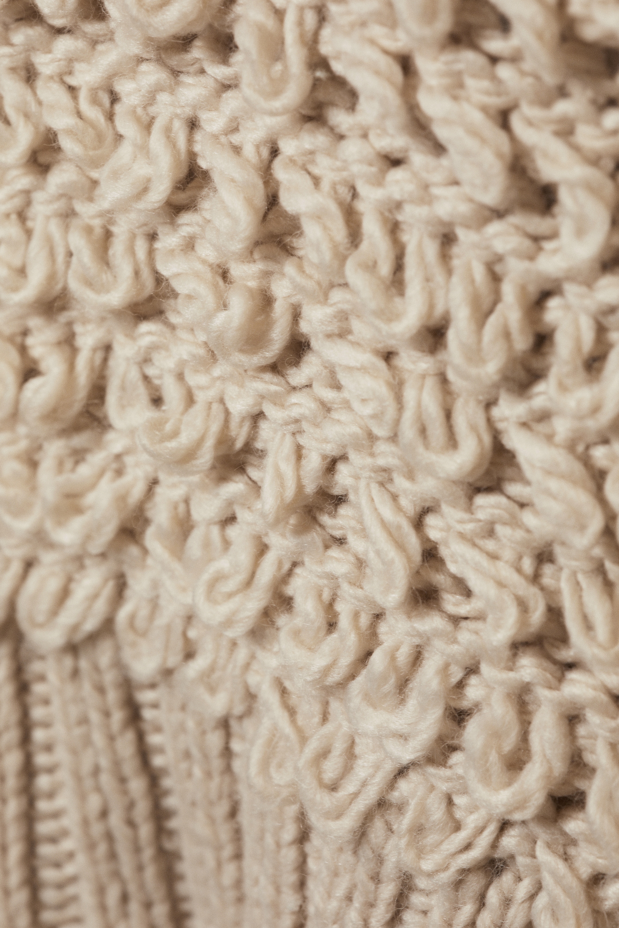 View larger image: A cream-colored knitted fabric is featuring a textured pattern of raised, looped stitches across its surface. A section of ribbed knitting is also visible along the bottom left.