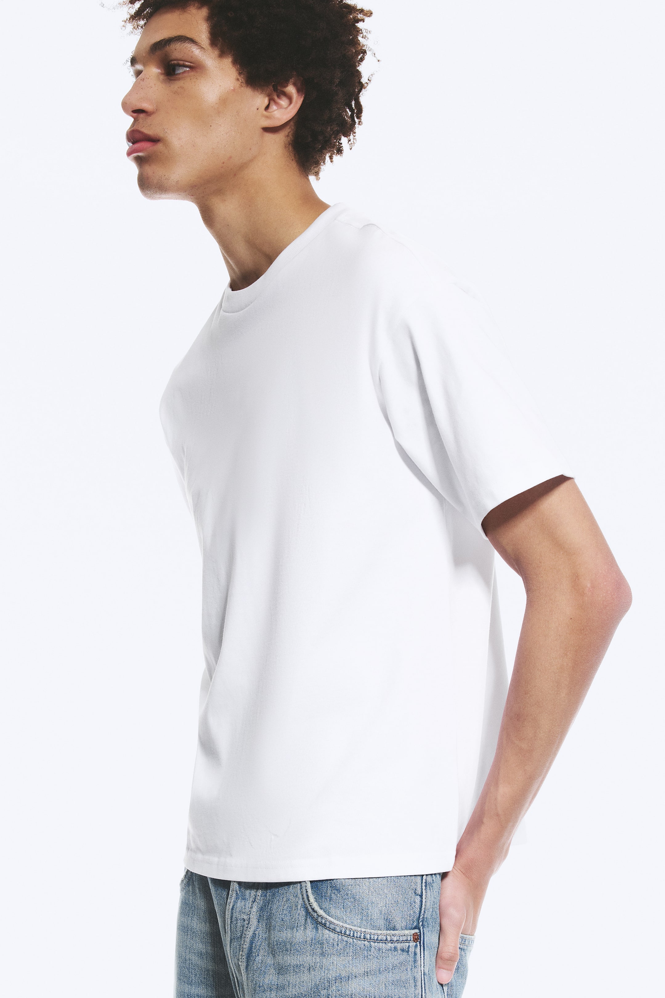 Se større billede: En hvid t-shirt præsenteres fra en sidevinkel, med rund hals, korte ærmer og en afslappet pasform, der falder over lyseblå jeans.