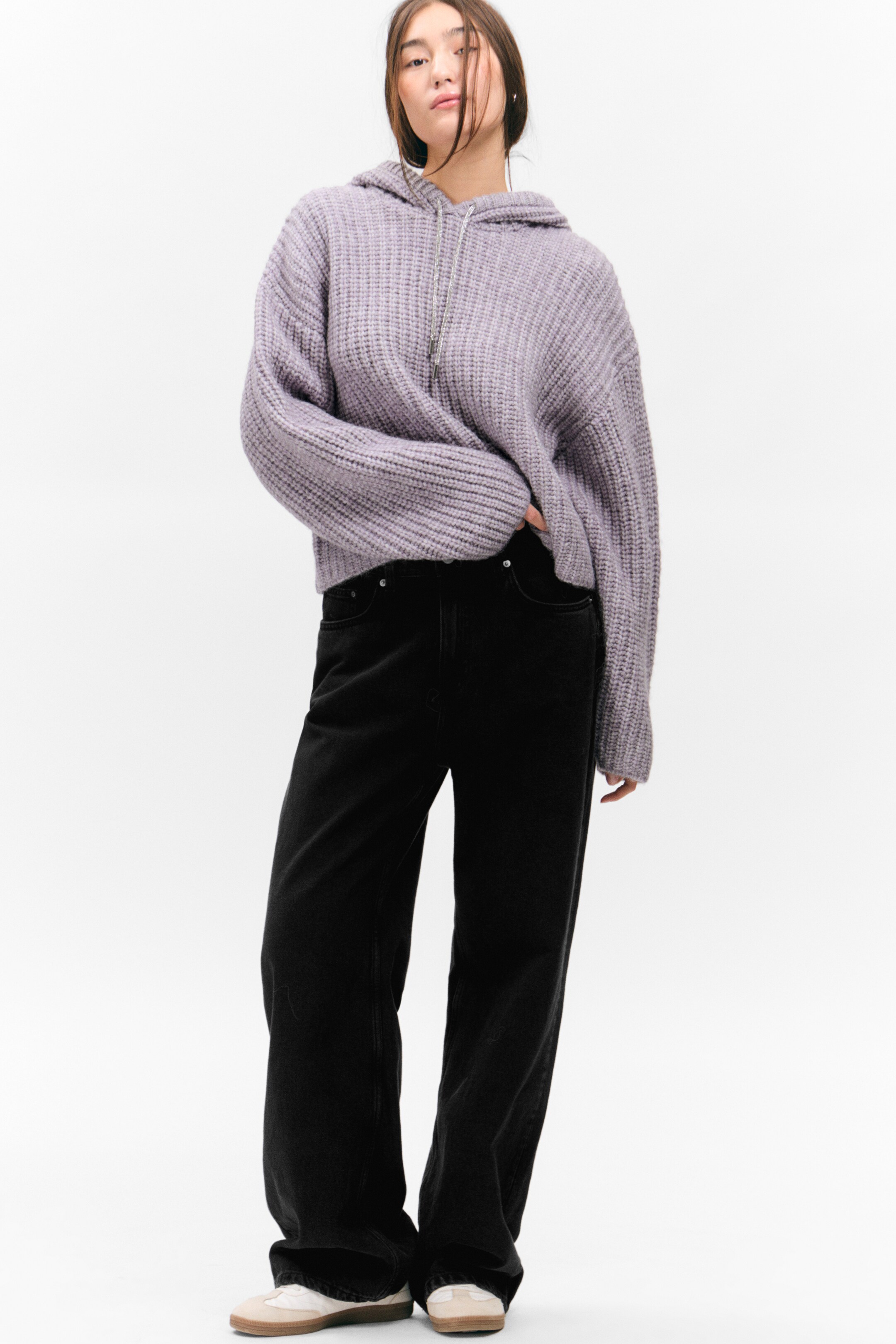 Größeres Bild anzeigen: Ein hellviolette, grob gestrickte Half-Zip-Kapuzenpullover wird aus einem leicht frontalen linken Winkel präsentiert und weist einen silberfarbenen Reißverschluss, eine Kapuze und eine lockere Passform auf. Der Kapuzenpullover wird über schwarzen, weit geschnittenen Jeans getragen, die mit Fronttaschen und einem hochgeschnittenen Design ausgestattet sind.