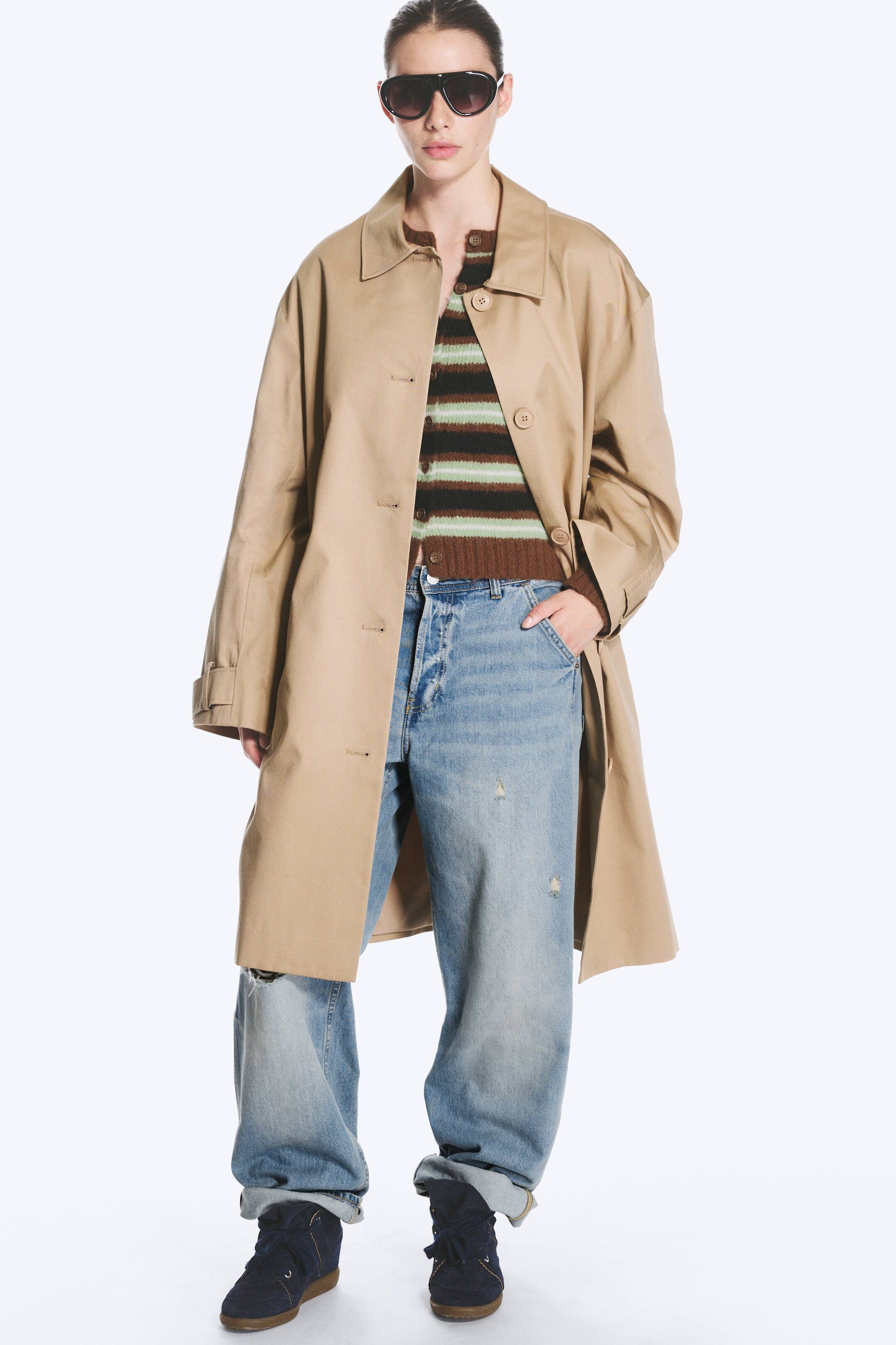 View larger image: A beige single-breasted car coat with a folded collar and dropped shoulders is worn open by the model. The coat is layered over a striped knit top and loose blue jeans, highlighting its relaxed fit and mid-length silhouette.