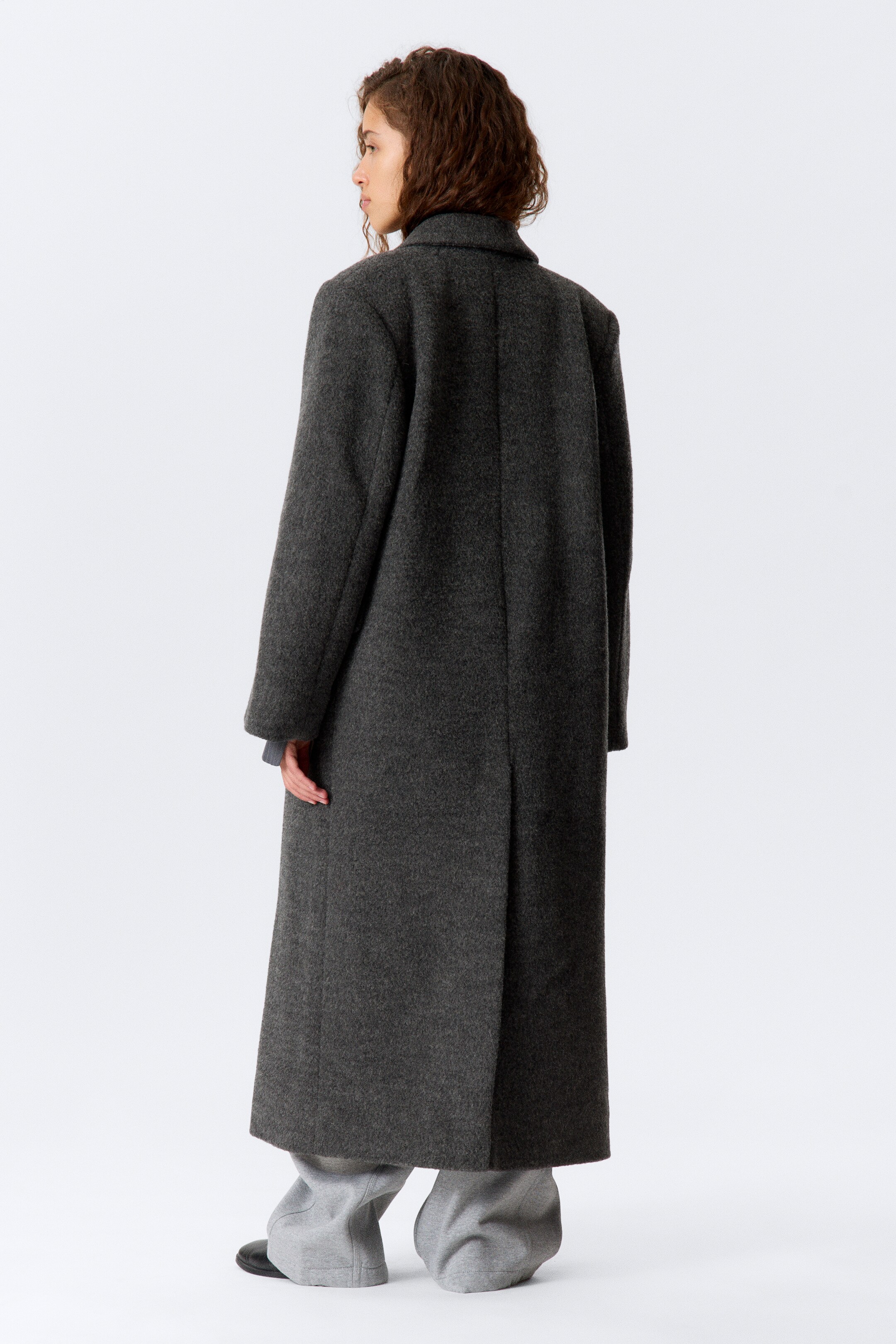 Grotere afbeelding bekijken: Een donkere grijze lange jas wordt vanachter gepresenteerd, lichtjes naar rechts gedraaid. Hij is voorzien van een geborsteld wol-mengsel stof, een brede kraag, en een oversized silhouet met brede schouders. De jas reikt net boven de enkels en toont een enkele rugsplit aan de zoom.