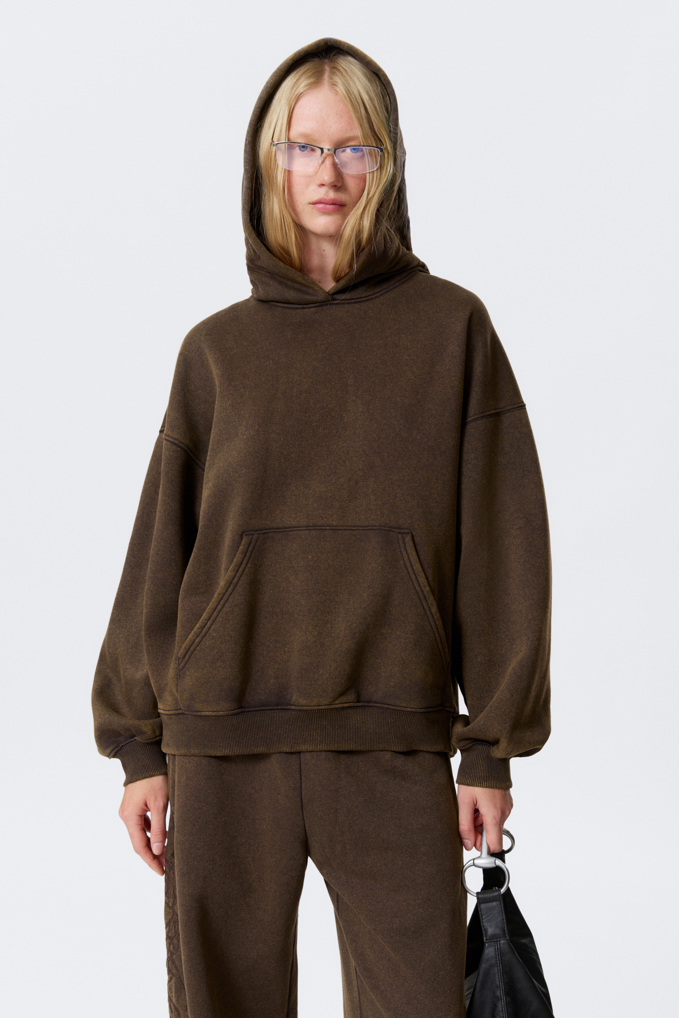 Visa större bild: En överdimensionerad, mörkbrun hoodie med en känguruficka, nedhasade axlar och ribbade muddar och fåll, bärs av modellen. Modellen har huvan uppfälld och hoodien är stylad med matchande mörkbruna träningsbyxor.