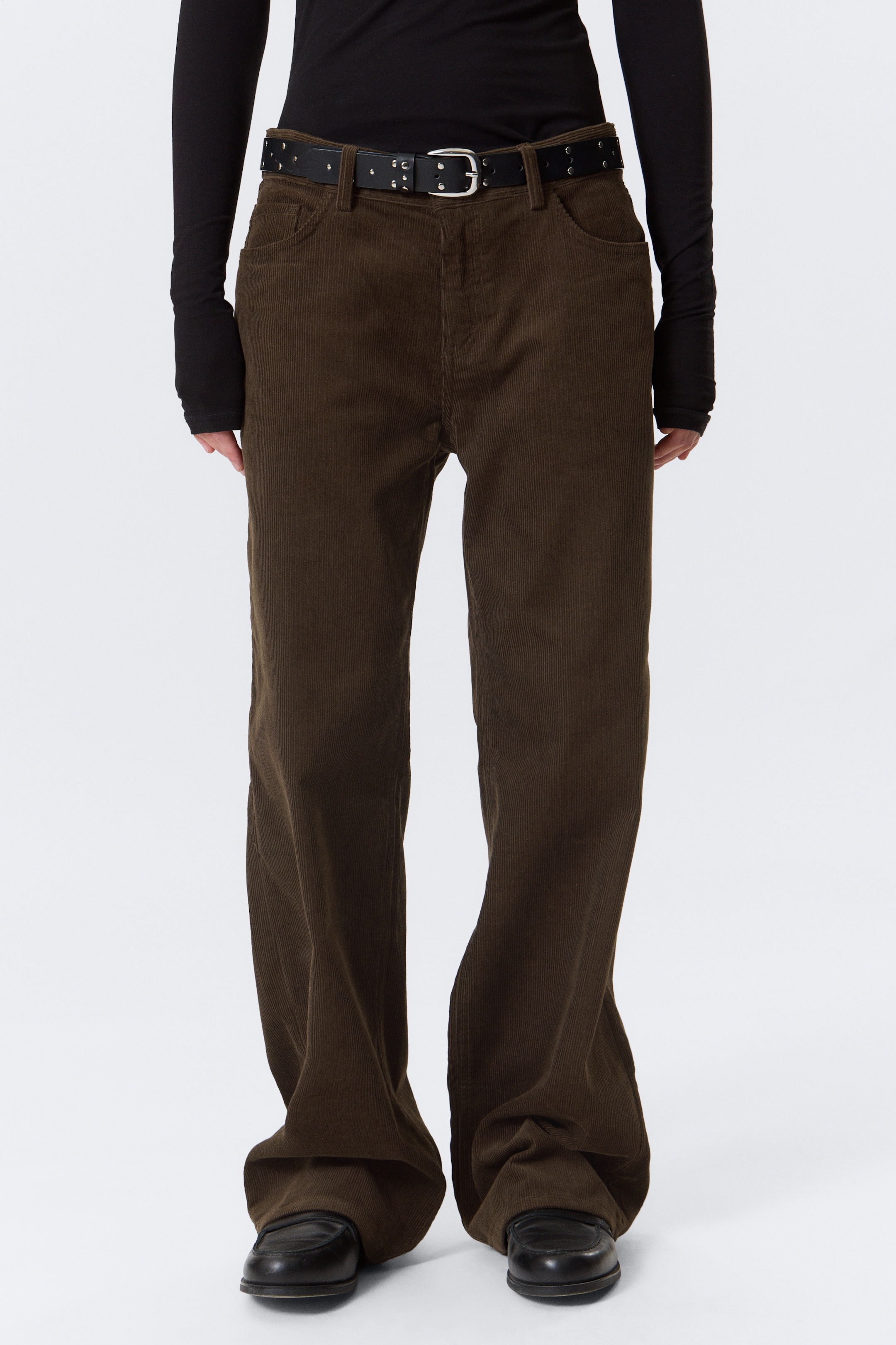 Visualizza immagine più grande: I pantaloni di velluto a coste marrone scuro sono presentati dalla parte anteriore, caratterizzati da un taglio a gamba larga con passanti per cintura visibili e tasche laterali che si appoggiano su scarpe nere. Sono indossati con una cintura in pelle nera con borchie.
