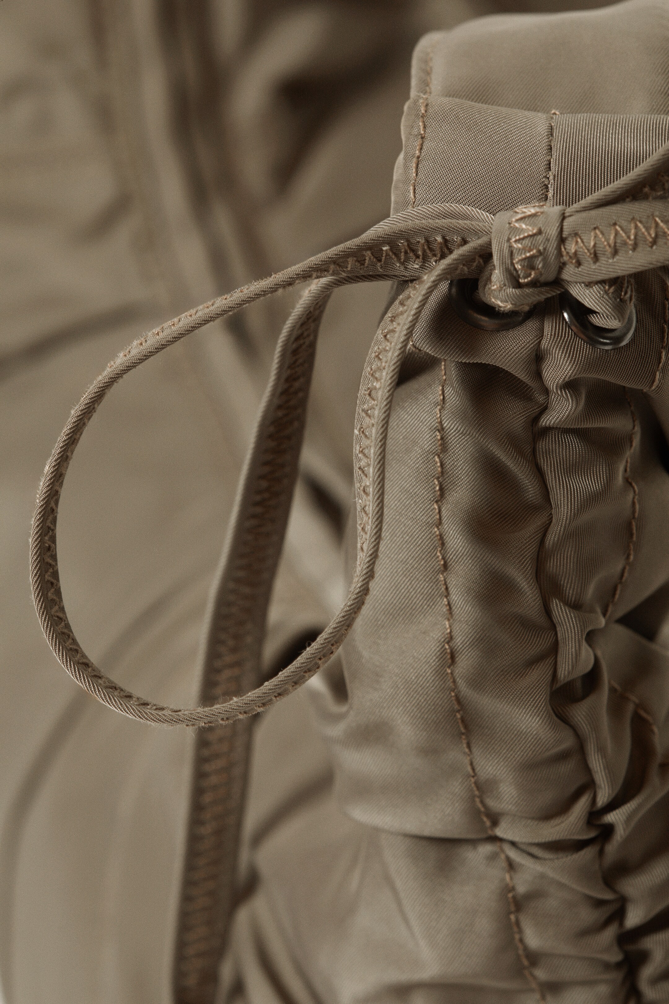 Vezi imaginea mai mare: Un detaliu de îmbrăcăminte de culoare taupe prezintă un șnur de material textil trecut prin două ocheți metalici. Materialul se adună în jurul șnurului, iar cusăturile drepte vizibile sunt prezente de-a lungul cusăturilor materialului, în timp ce o cusătură în zig-zag este vizibilă pe șnurul propriu-zis.