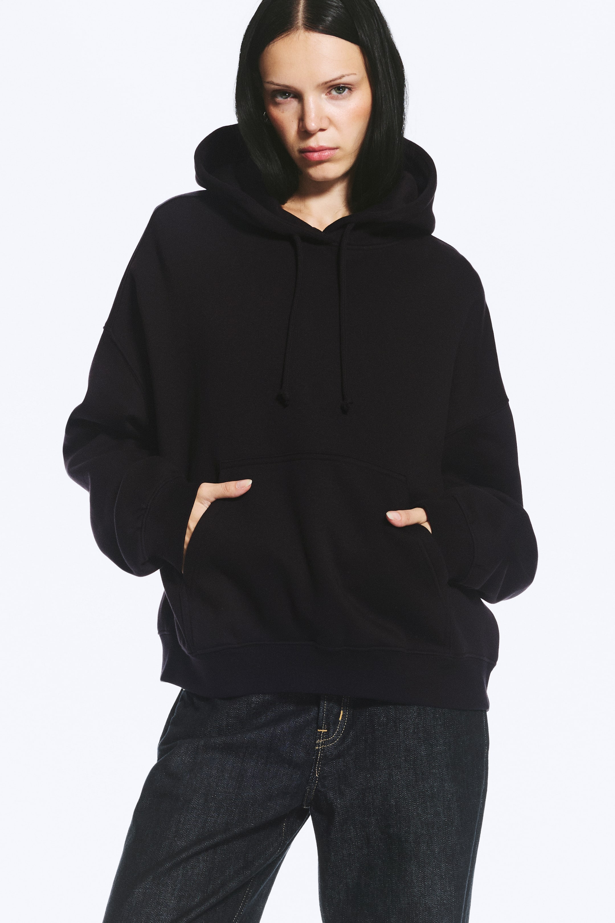 Ver imagen más grande: La modelo lleva una sudadera con capucha de corte regular y color negro, con hombros caídos, mangas largas y acabados acanalados. Presenta una capucha amplia con cordón y un gran bolsillo canguro, en el que la modelo está introduciendo ambas manos.