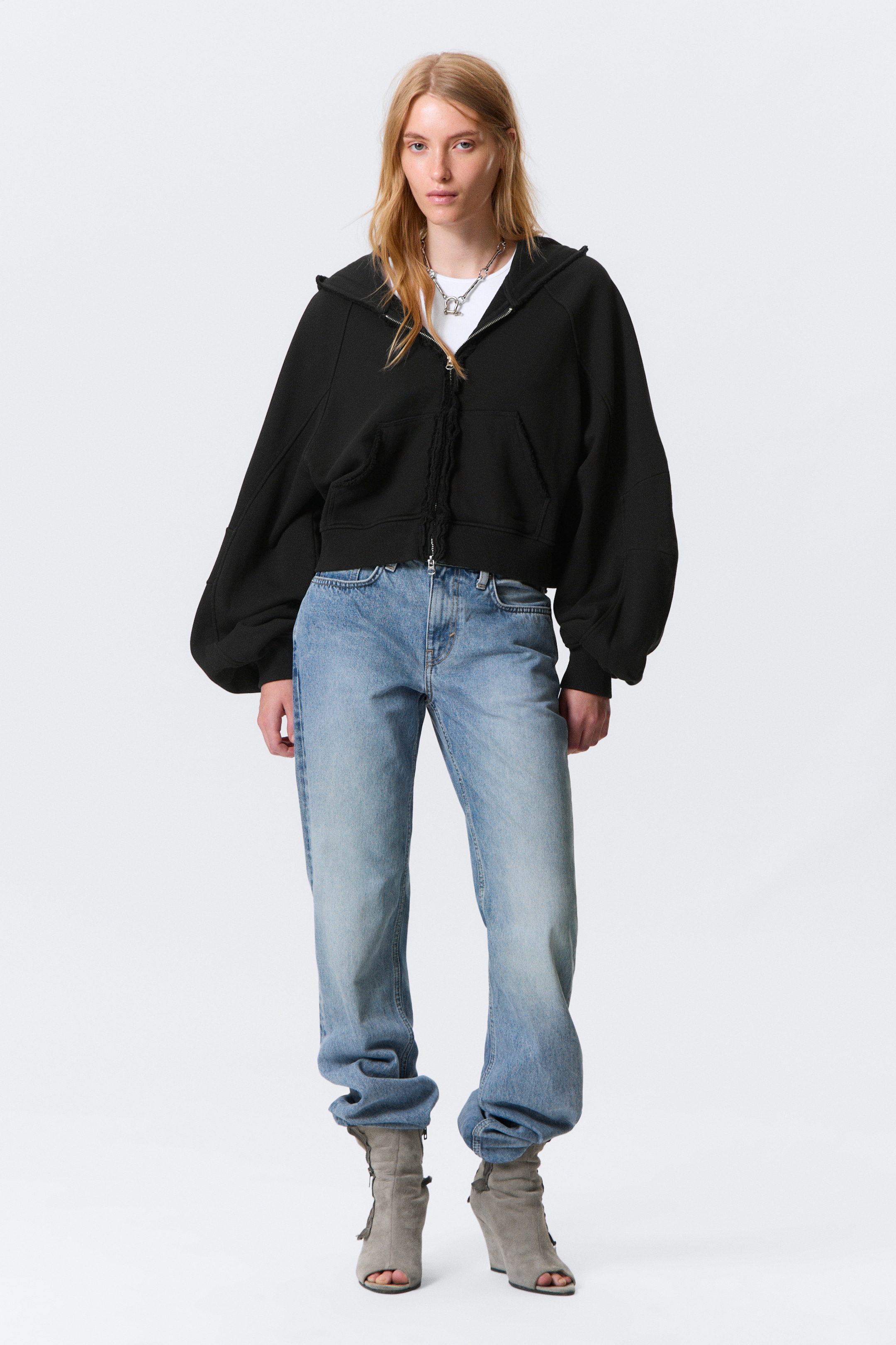 Größeres Bild ansehen: Ein schwarzer Hoodie mit durchgehendem Reißverschluss wird von vorne präsentiert, mit einer verkürzten, übergroßen Silhouette, voluminösen Ärmeln und gerafften Bündchen. Er zeigt einen durchgehenden Reißverschluss, eine Kängurutasche mit rohen Kanten und einen Saum mit rohen Kanten, während ein weißes T-Shirt darunter geschichtet ist.