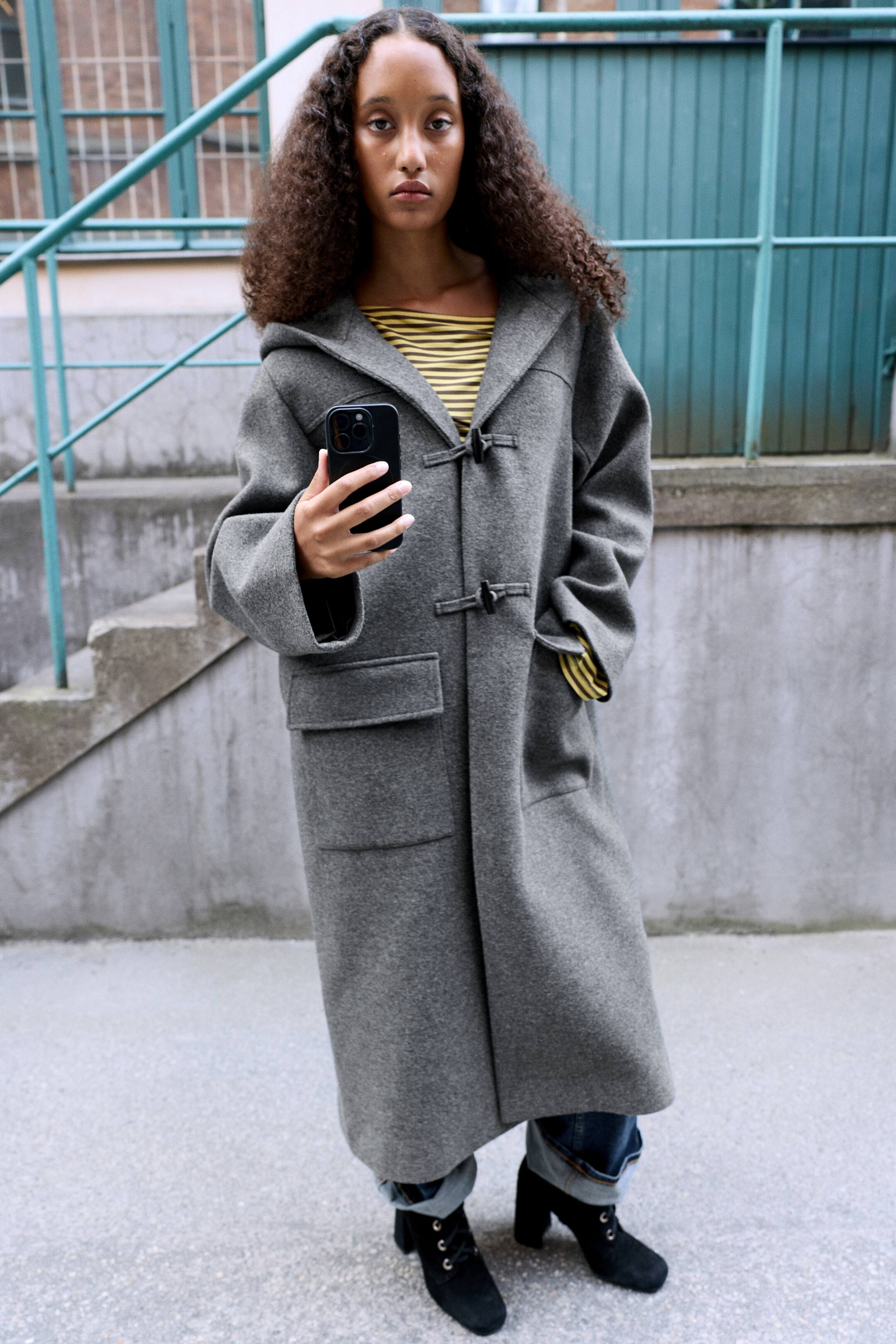 View larger image: A grey A-line duffle coat featuring a hood, toggle fastenings, and large front pockets is being worn by the model. The coat is layered over a yellow-striped top and cuffed jeans, while the model holds a smartphone in one hand and rests the other in a pocket.