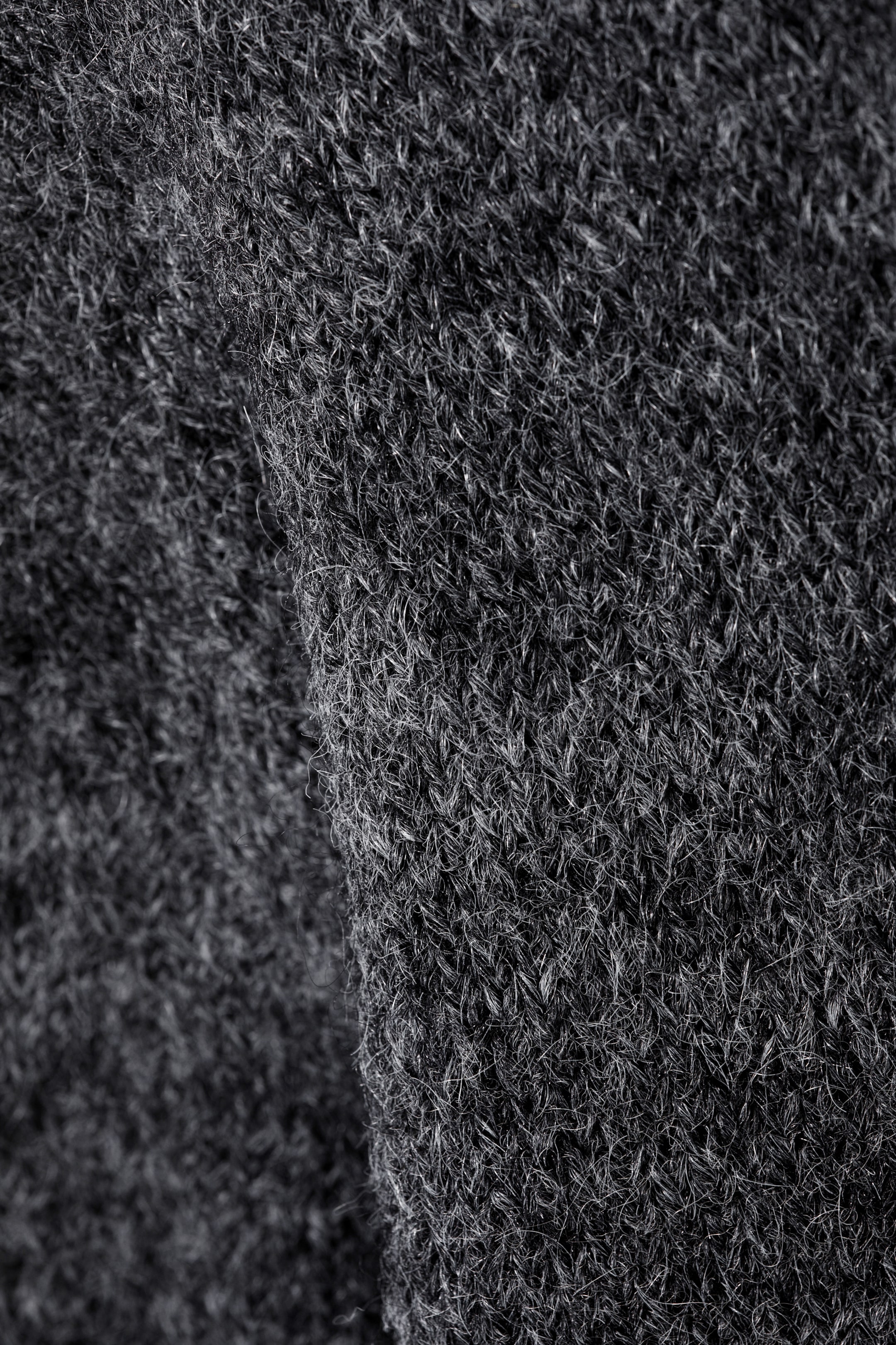 Grösseres Bild ansehen: Eine Nahaufnahme zeigt den weichen, dunkelgrauen Strickstoff eines Pullovers. Die Textur erscheint leicht flauschig, mit sichtbaren einzelnen Fasern, charakteristisch für eine Wollmischung.