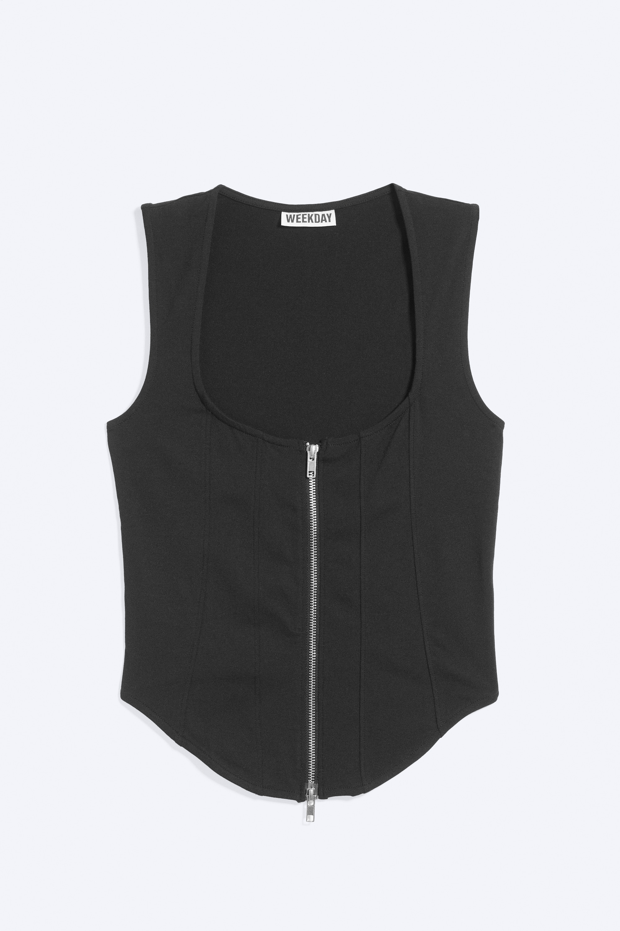 La modella indossa un corsetto nero aderente con zip, caratterizzato da scollo rotondo, cuciture verticali e orlo curvo a punta. Il top senza maniche presenta una vistosa cerniera frontale argentata a tutta lunghezza ed è abbinato a jeans blu a vita bassa.