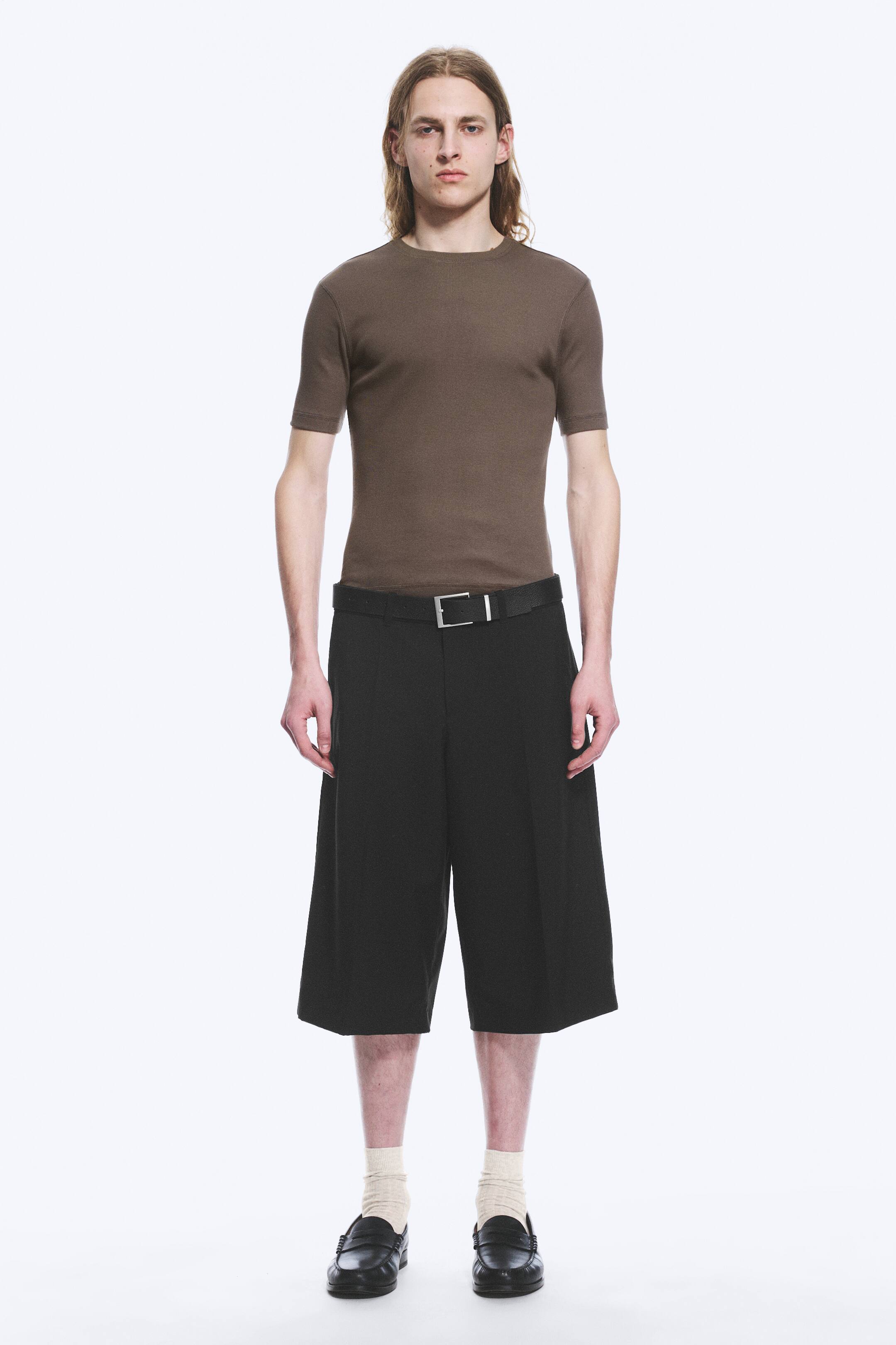 View larger image: The model is wearing a brown fitted short-sleeve t-shirt. Accompanying this, the model has on black wide-leg shorts, fastened with a black belt featuring a silver rectangular buckle, and black loafers paired with light cream socks.