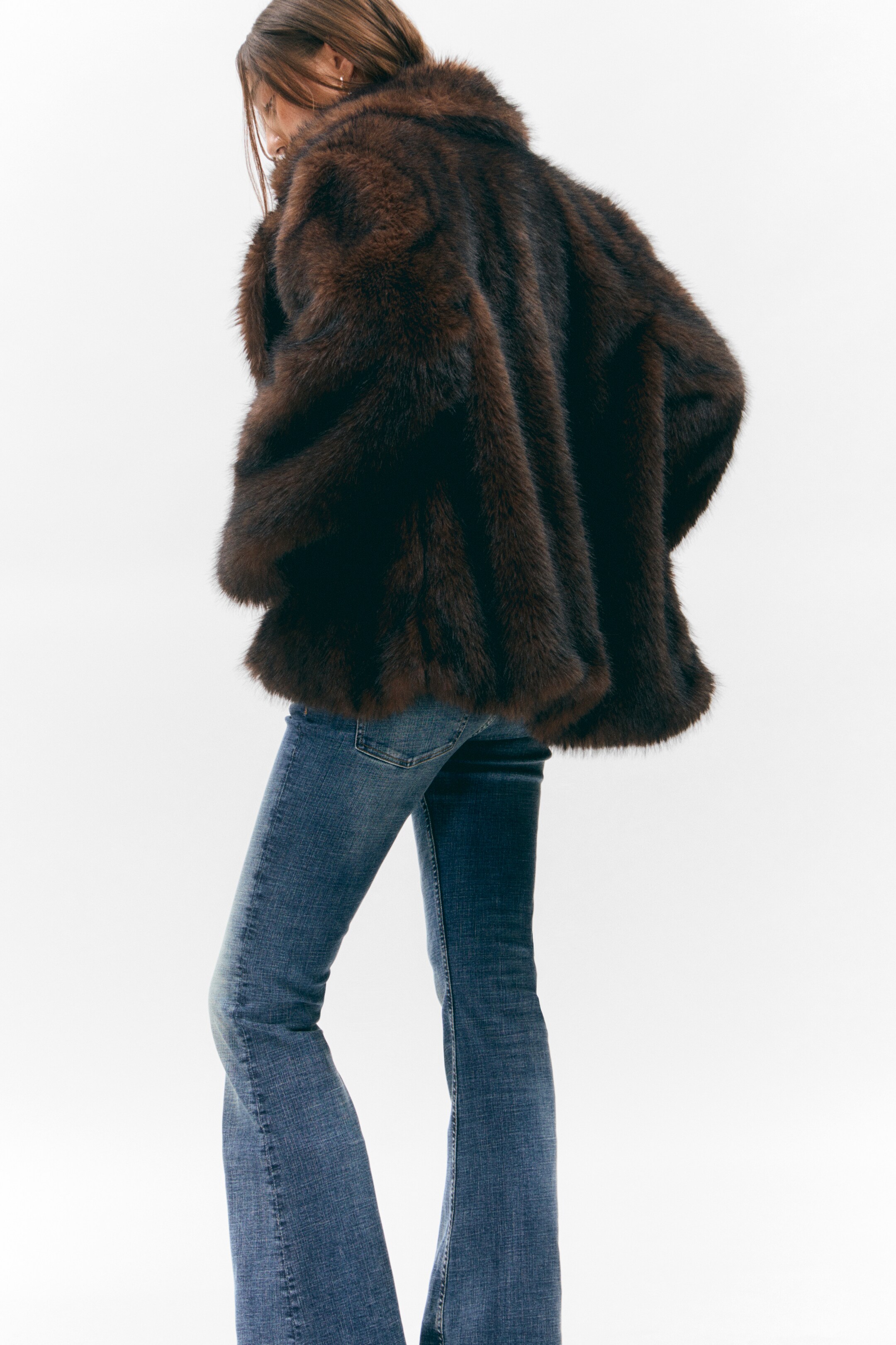 Agrandir l'image: Un manteau en fausse fourrure brun volumineux et multi-tons, doté d'un large col debout, est porté par l'arrière et légèrement vers la droite. Il est superposé à un jean en denim bleu avec une jambe évasée.