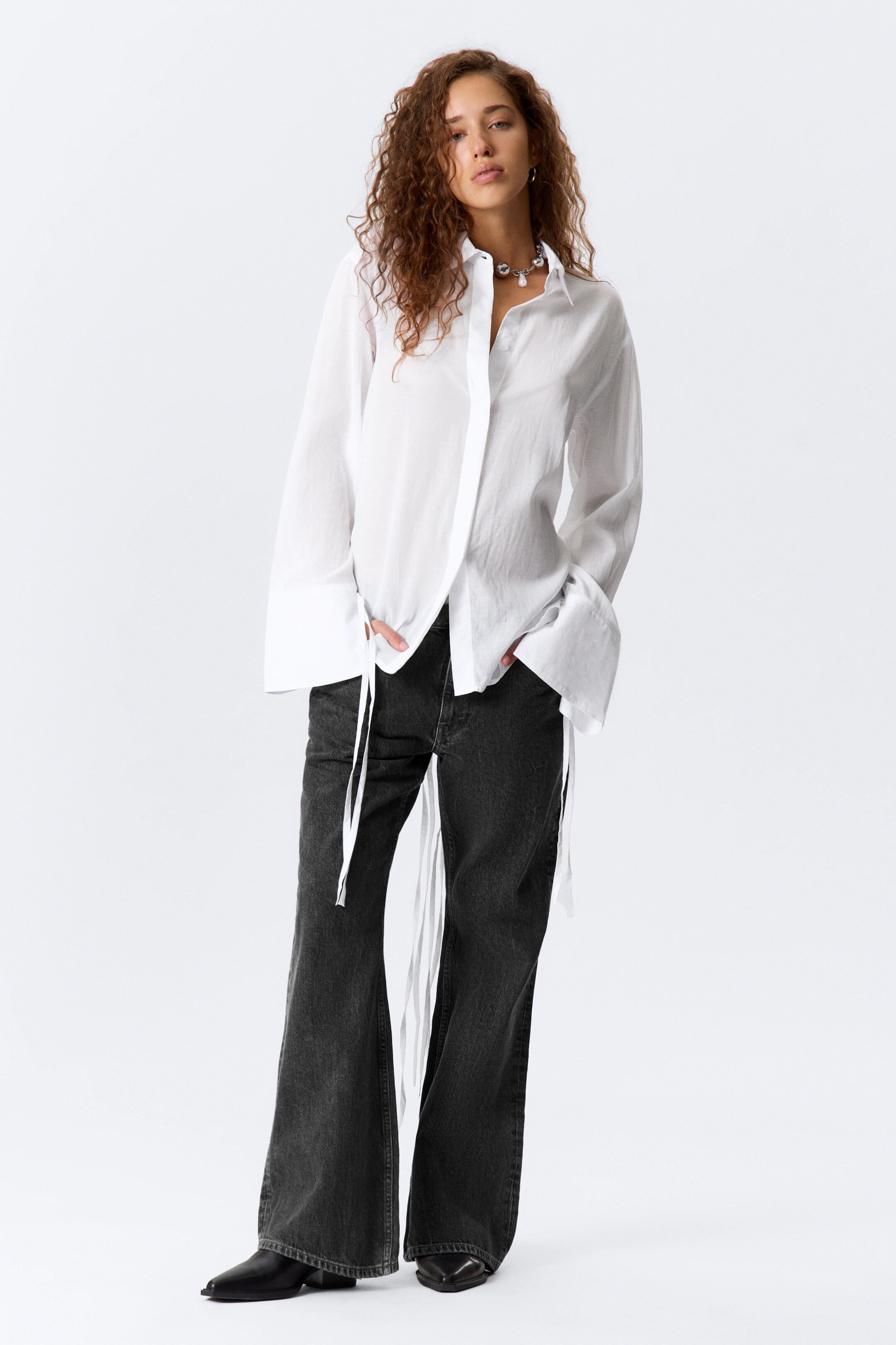 Ver imagem maior: Uma camisa branca oversized, com gola pontiaguda, mangas compridas e largas com punhos dobrados profundos e laços longos que se estendem das costuras laterais, está a ser apresentada com jeans em tom cinza escuro e de perna larga.