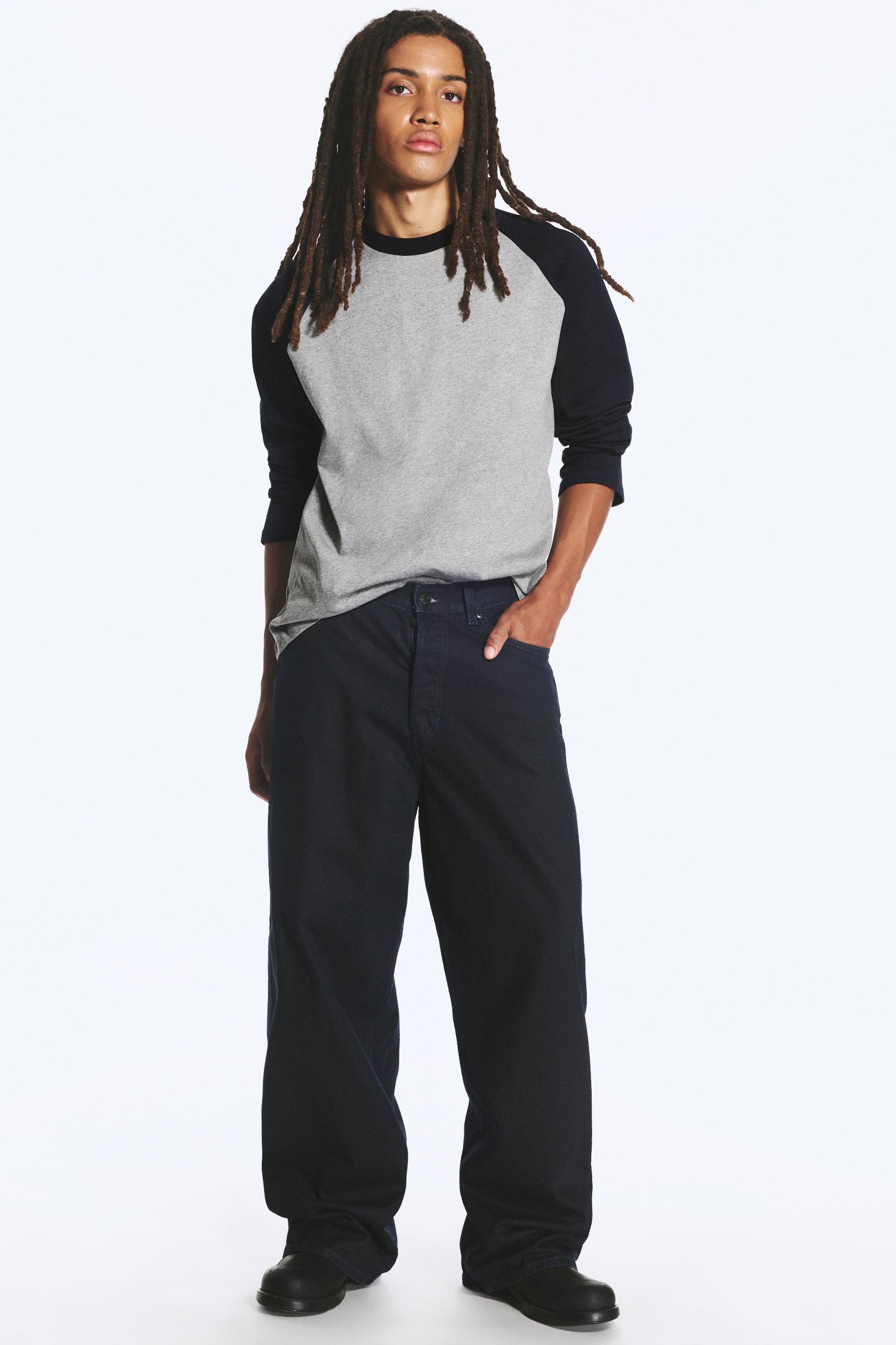 Agrandir l'image: Le mannequin porte un jean enduit bleu indigo foncé à jambe ample et baggy, avec une main posée dans la poche avant. Ce jean taille mi-haute est associé à un t-shirt raglan gris et noir et à des chaussures noires.