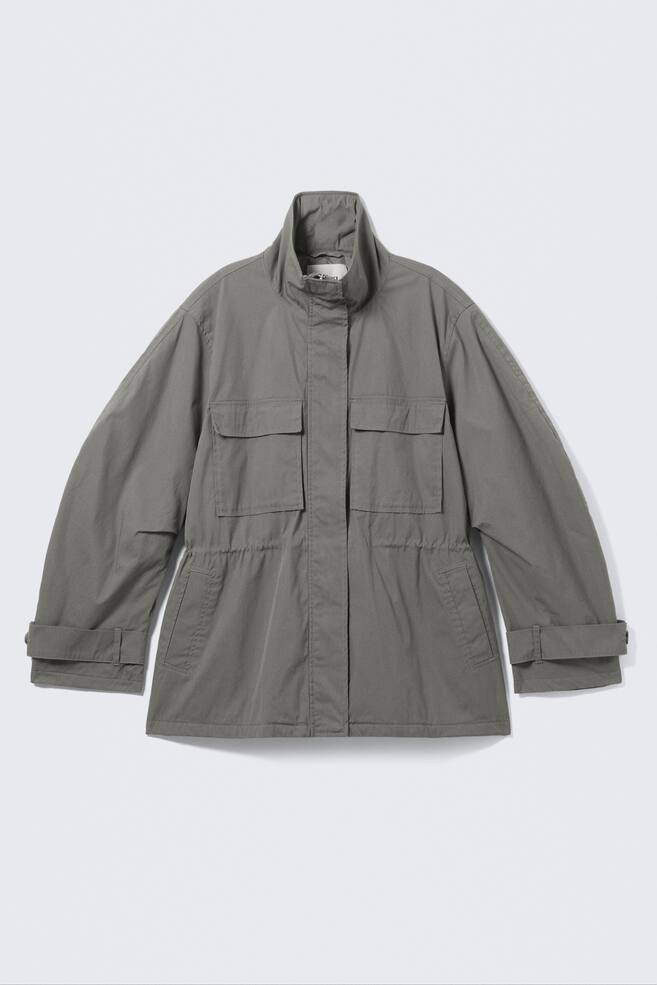 Una giacca grigio-verde presenta il suo FRONTE, caratterizzata da un collo alto a fascia e una chiusura frontale nascosta. Mostra due ampie tasche sul petto con patta, due tasche laterali inclinate, un cordoncino elastico che stringe la vita, linguette regolabili sui polsini delle maniche arrotolate e un orlo dritto. - 1