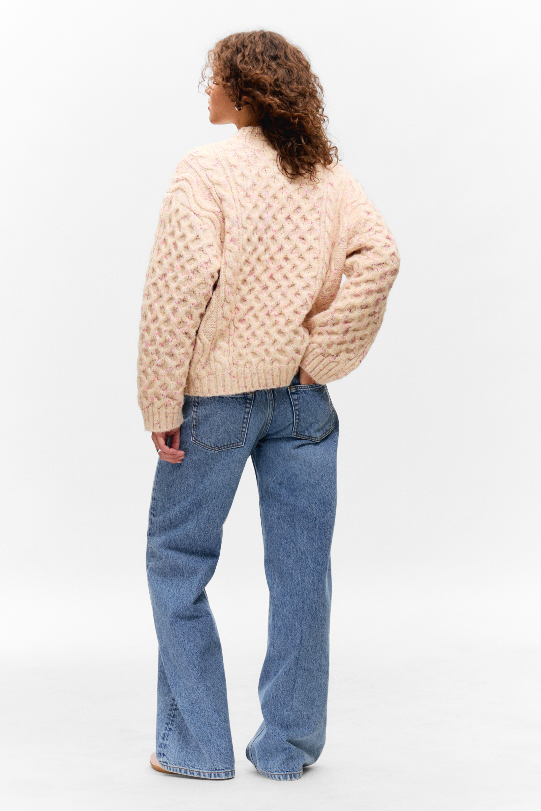 Se større billede: En cremefarvet, chunky kabelstrikket sweater med lyserøde pletter bæres, med ryggen vendt mod beskueren. Den har fremtrædende diamant- og lodrette kabelstrikmønstre, voluminøse ærmer, en afslappet pasform og en ribbet kant, og den er parret med mellemblå, afslappet pasform, bredbenede jeans.