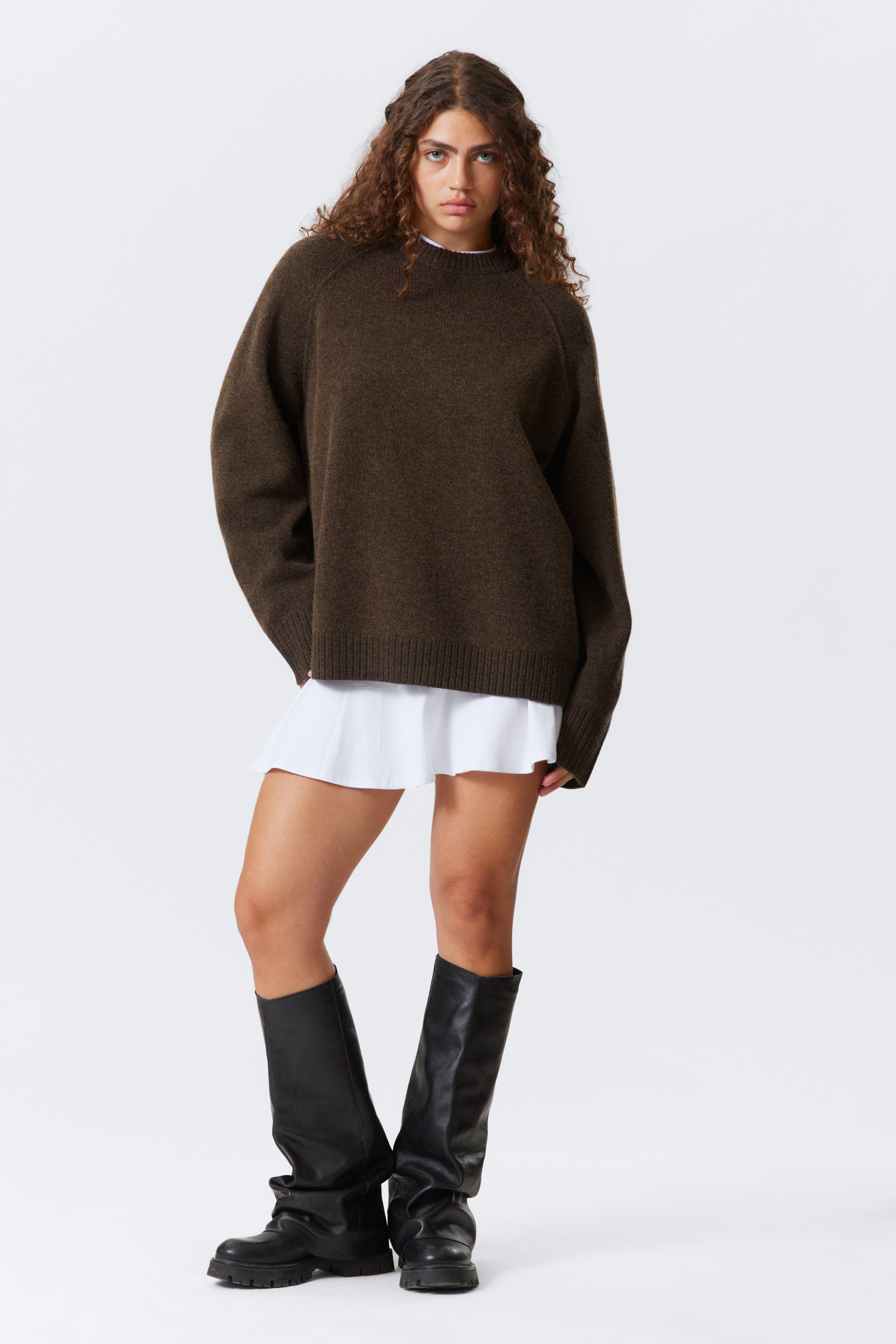 Größeres Bild anzeigen: Ein dunkelbrauner Strickpullover mit lockerem Schnitt und Raglanärmeln wird vom Model getragen. Er zeigt breite gerippte Abschlüsse an den Bündchen, am Saum und am Halsausschnitt und wird über einem weißen Kleidungsstück getragen.