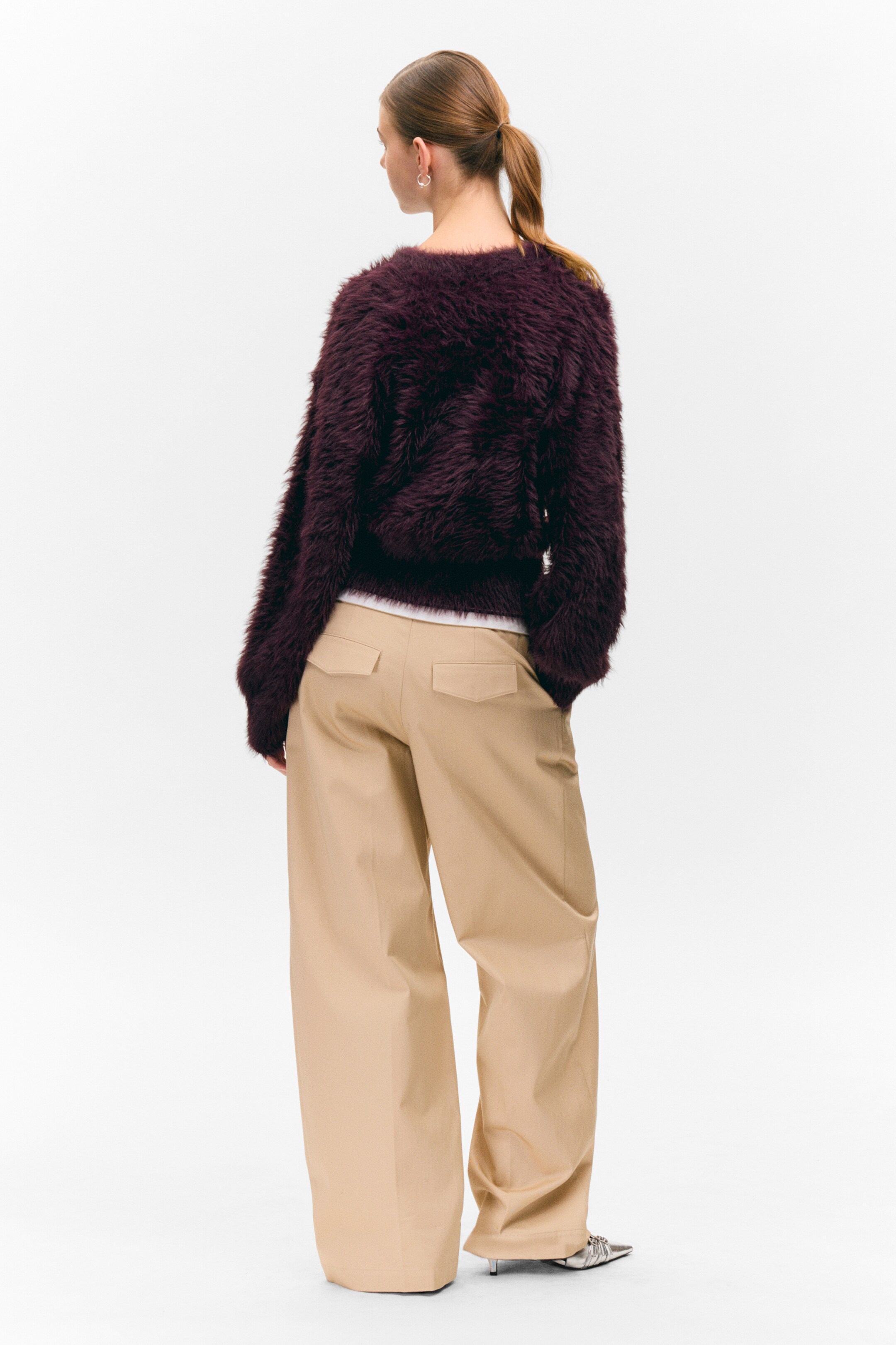Se større billede: En mørk lilla, lurvet strikket sweater vises fra bagsiden, hvilket fremhæver dens voluminøse tekstur, parret med beige bukser med brede ben og metalliske spidse sko. Bukserne har to baglommer med klap og en afslappet, flydende pasform, med et hvidt lag synligt under sweateren.