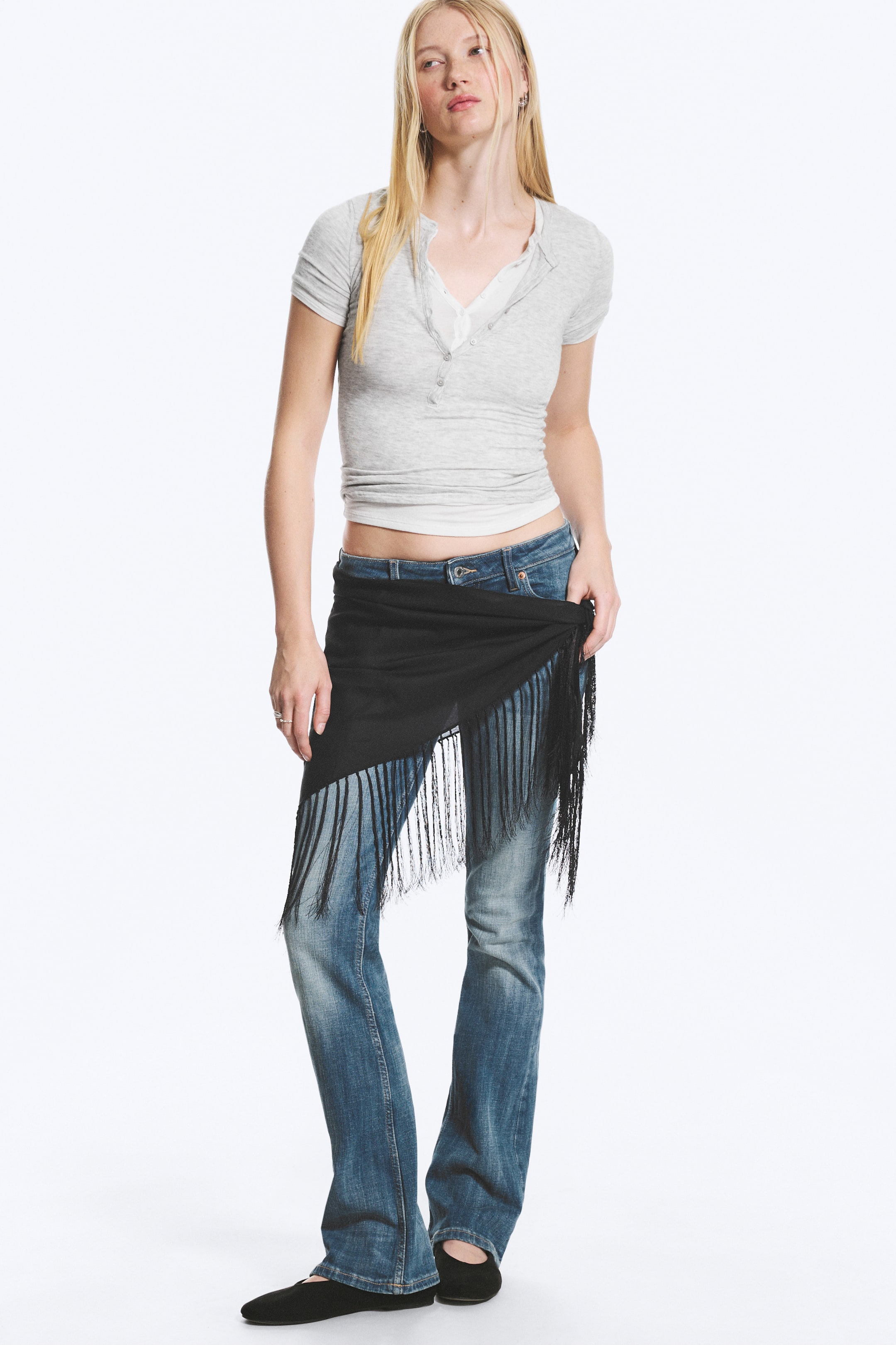 Visualizza immagine più grande: Un pareo nero con frange è indossato dalla modella, drappeggiato intorno alla vita sopra un paio di jeans blu. La modella tiene una sezione del pareo con una mano.
