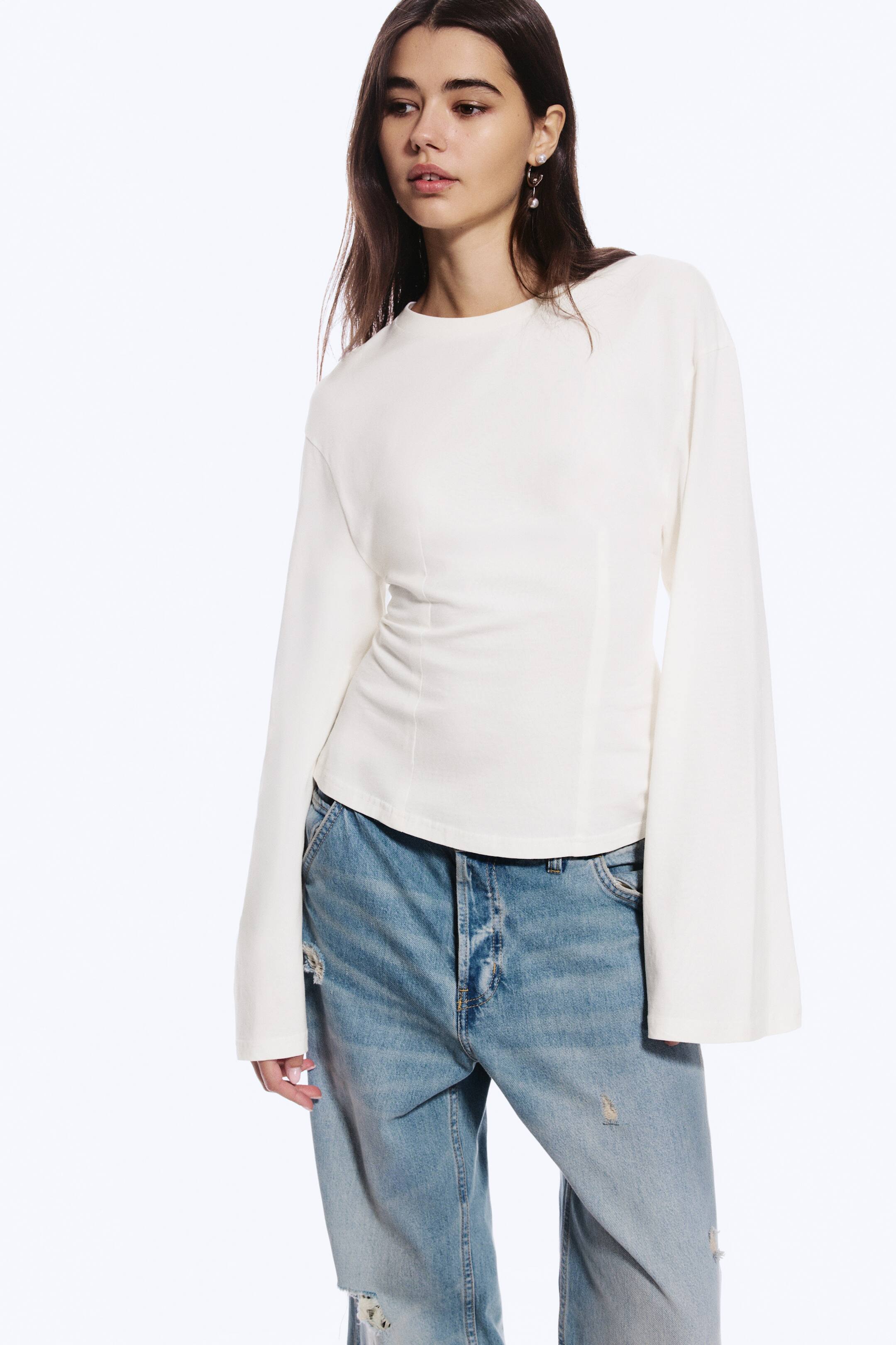 Grösseres Bild ansehen: Das Model trägt ein weißes Langarm-Top mit Rundhalsausschnitt, einer taillierten Passform mit vertikalen Formnähten und langen, ausgestellten Ärmeln. Das Top ist mit verwaschenen Blue Jeans kombiniert.