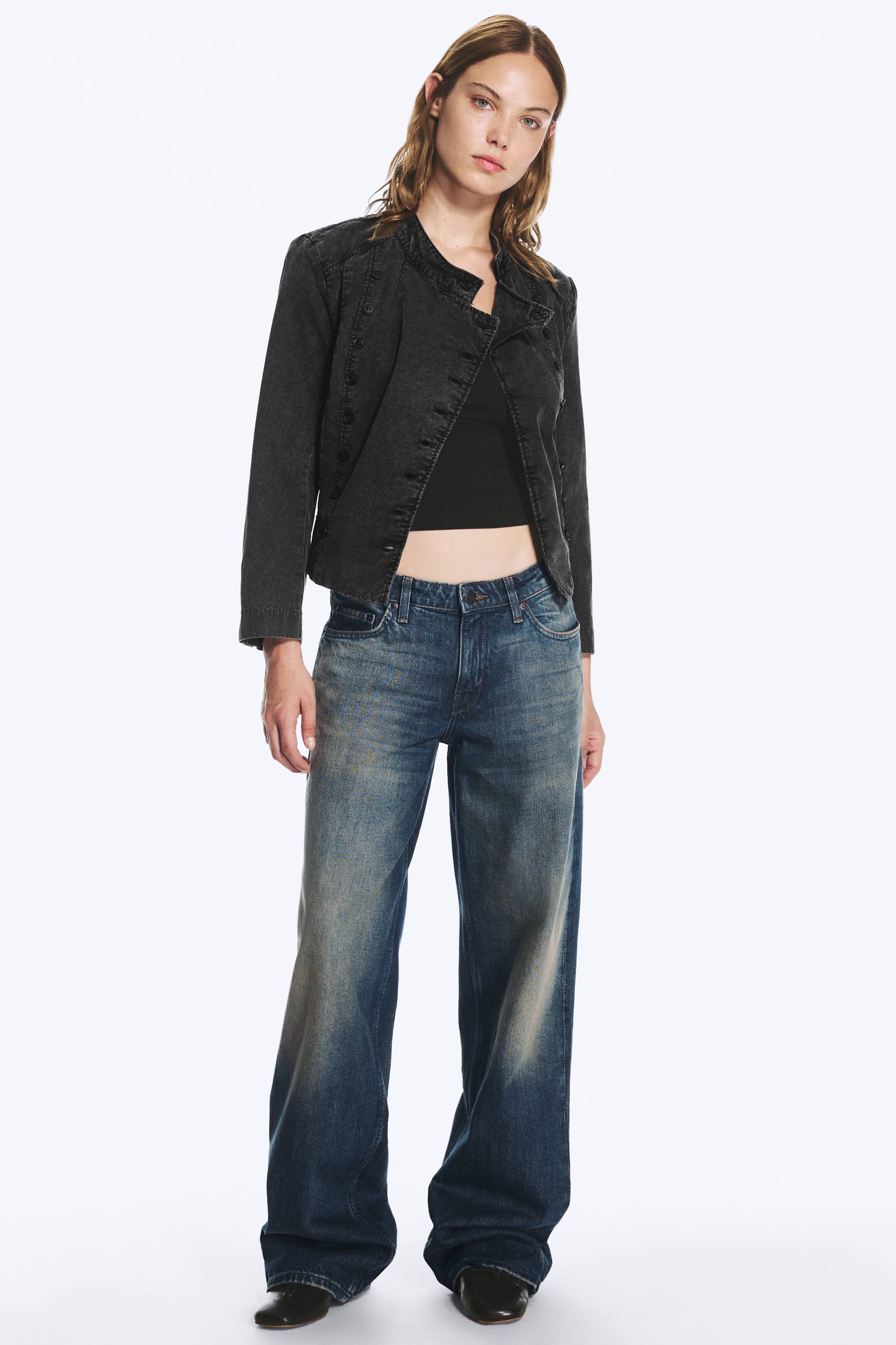 Agrandir l'image: Une paire de jeans bleus taille basse à jambes larges, avec un délavage effet usé, est portée par le mannequin. Ce jean ample et long est associé à un top court noir, une veste boutonnée gris foncé et des chaussures noires.