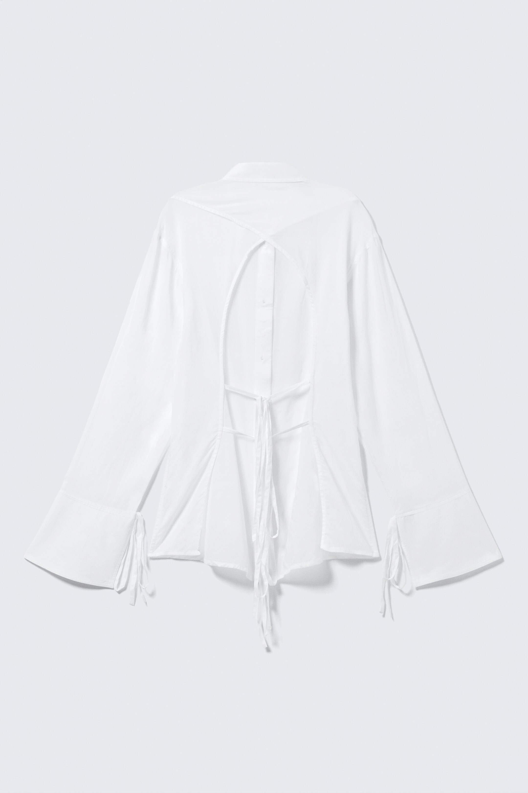 Ver imagem maior: Uma camisa branca está a mostrar as costas, apresentando uma gola e um recorte em forma de V na parte superior das costas, revelando uma abas de botões. Um fecho horizontal com laço está a apertar a cintura, com o tecido a franzir e a alargar abaixo, e as mangas largas terminam com detalhes de laço nos punhos.