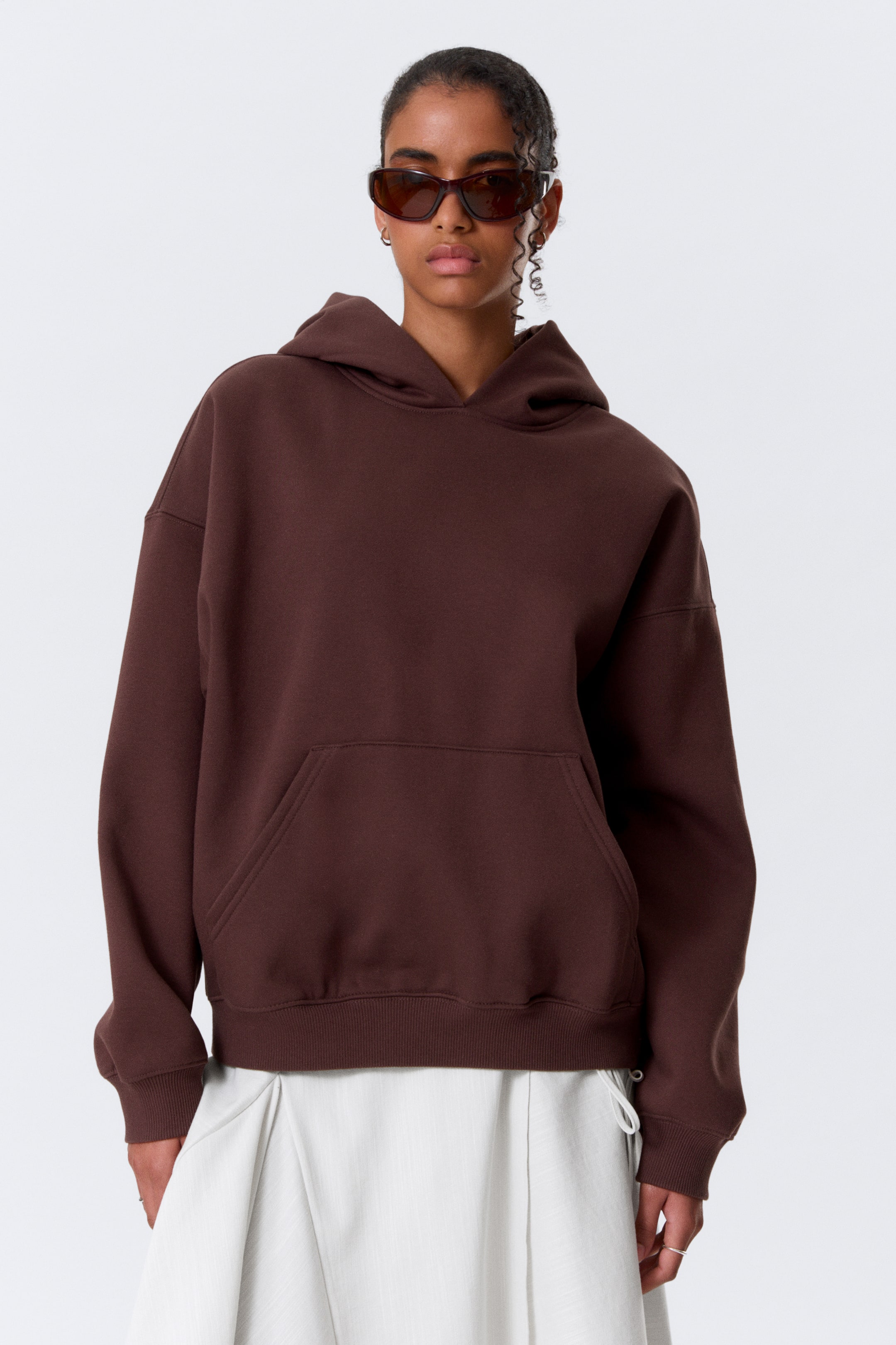 Προβολή μεγαλύτερης εικόνας: Μια καφέ φαρδιά κουκούλα, με χαλαρή σιλουέτα και χαμηλωμένους ώμους, μια τσέπη τύπου καγκουρό, μια μεγάλη, ευρύχωρη κουκούλα χωρίς κορδόνι και ριγέ στρίφωμα, φοριέται από το μοντέλο. Το μοντέλο το συνδυάζει με ανοιχτόχρωμο παντελόνι με πιέτες.