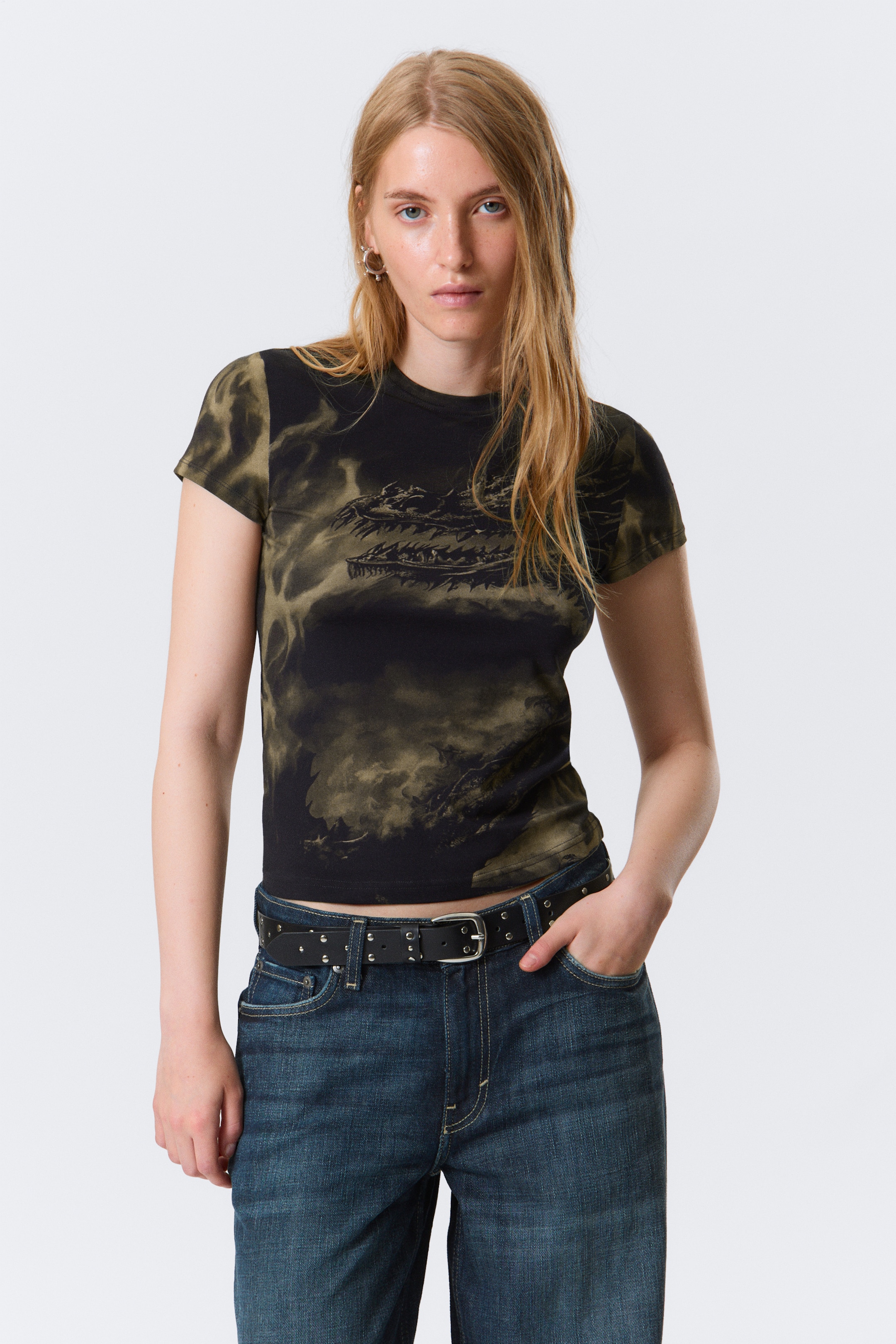 Größeres Bild anzeigen: Ein kurzärmeliges T-Shirt in einem schwarz-olivgrünen marmorierten Muster zeigt auf der Vorderseite eine dunkle Drachengrafik. Das Rundhals-Oberteil ist mit einer eng anliegenden, kurzen Silhouette geschnitten und wird mit dunkelblauen Jeans und einem schwarzen, mit Nieten besetzten Gürtel getragen.