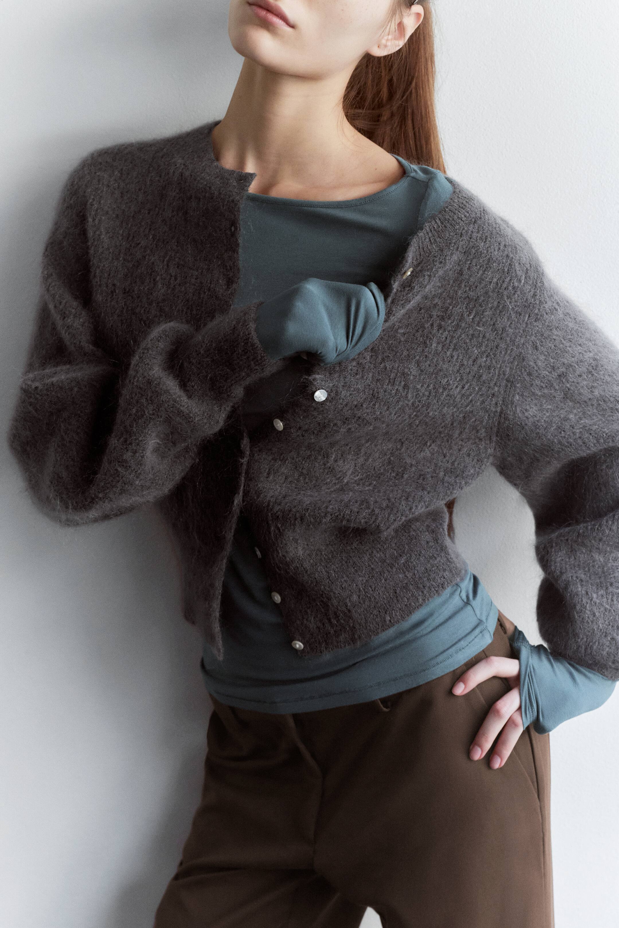 Grösseres Bild ansehen: Eine graue, gebürstete Strickjacke aus Mohair-Mischung wird vom Model über einem petrolfarbenen Langarmtop und braunen Hosen getragen. Das Model richtet die Vorderseite der teilweise geknöpften Strickjacke und betont damit ihren runden Ausschnitt, die kleinen weissen Knöpfe sowie die langen Ärmel mit Rippbündchen.
