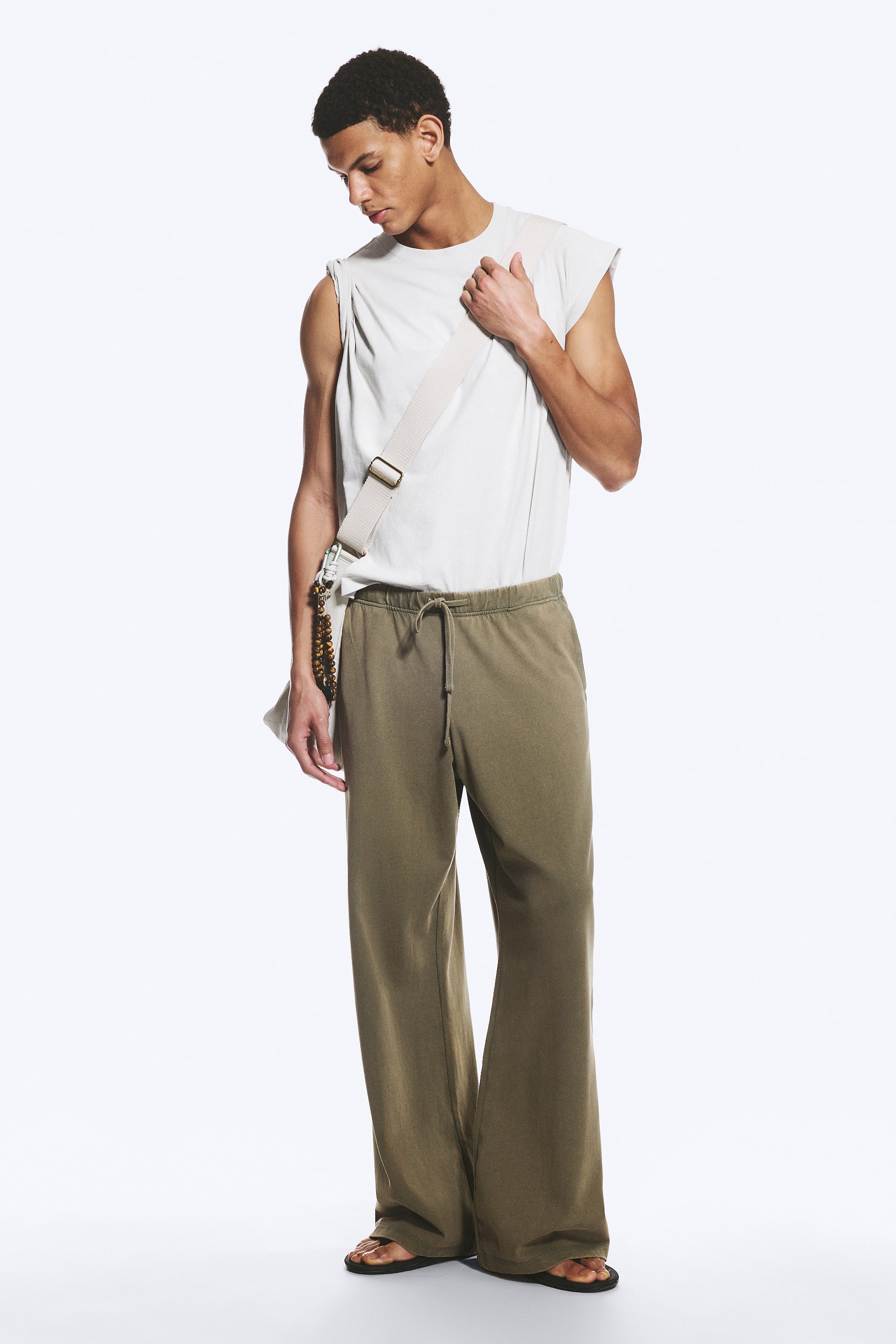 Se større billede: En off-white ærmeløs muscle tee med rund hals er stylet med olivengrønne sweatpants med brede ben, der har en snøre i taljen og en afslappet pasform. En let canvas crossbody-taske med en bred rem og dekorative perler bæres, hvilket fuldender det afslappede ensemble.