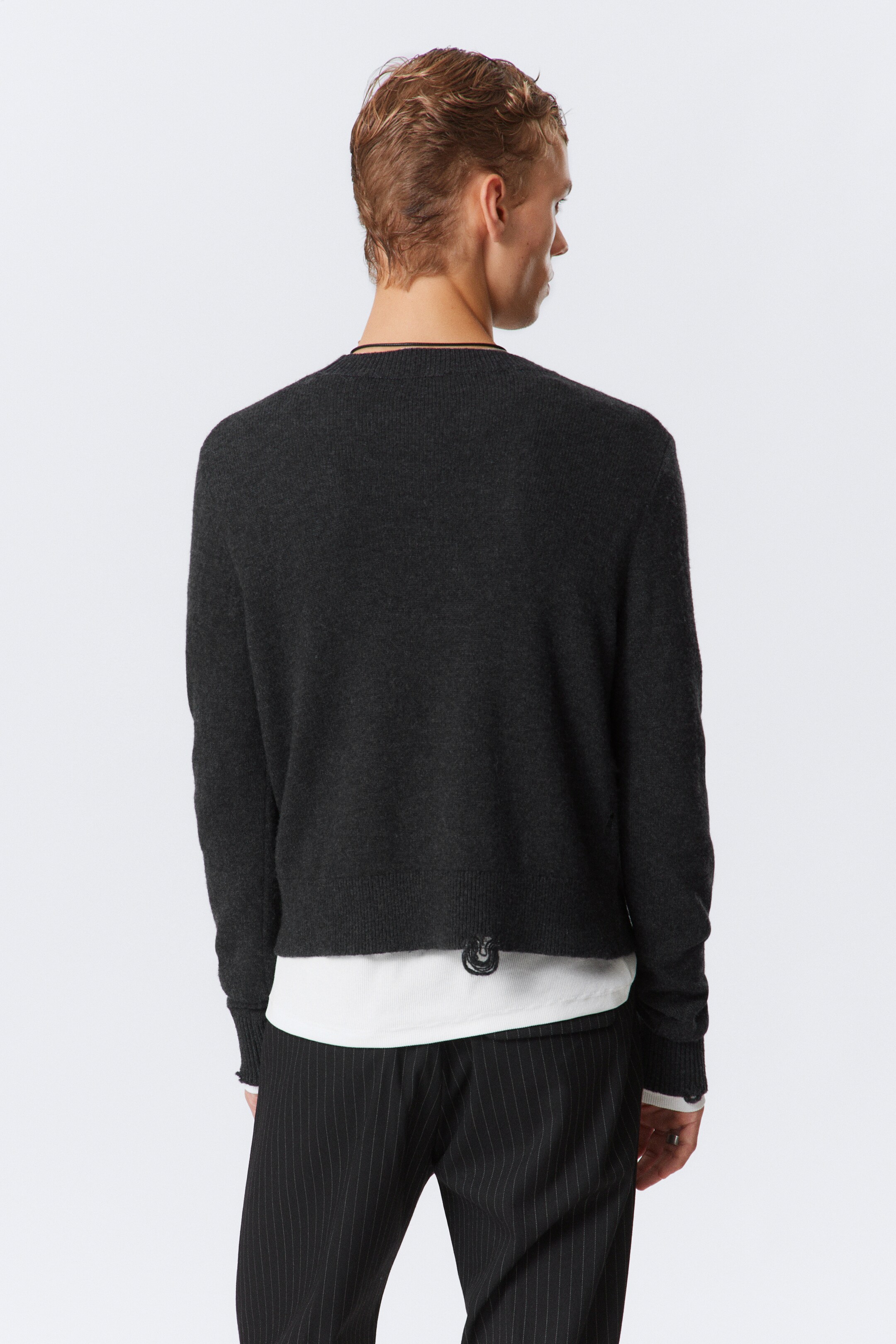 View larger image: A dark gray knitted sweater is being shown from the back, featuring a crew neck, long sleeves with ribbed cuffs, and a ribbed hem. It is being layered over a white long-sleeved shirt, which is visible at the neckline, cuffs, and hem, and a small black loop detail is appearing at the sweater's lower left back hem.