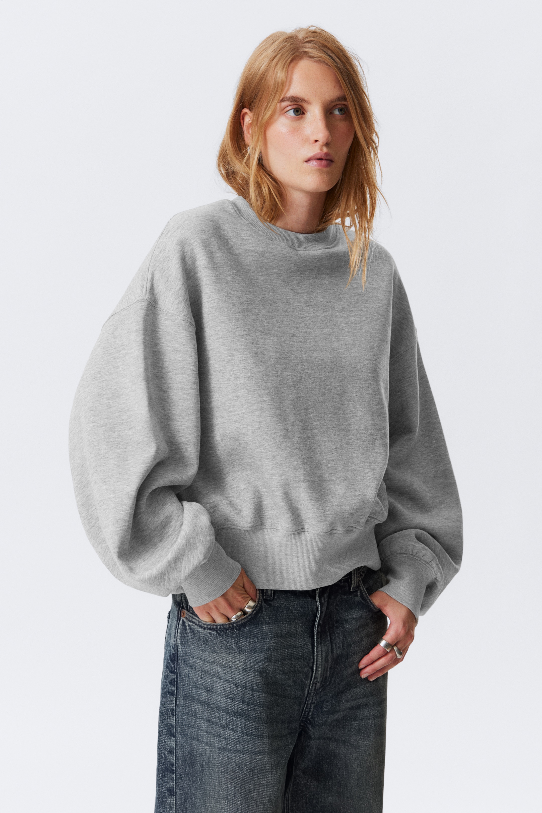 Größeres Bild anzeigen: Ein graues Sweatshirt mit Rundhalsausschnitt wird getragen, das eine übergroße Passform mit voluminösen langen Ärmeln und gerippten Bündchen aufweist. Sein gerippter Saum ruht auf der Taille von Jeans in dunkler Waschung.