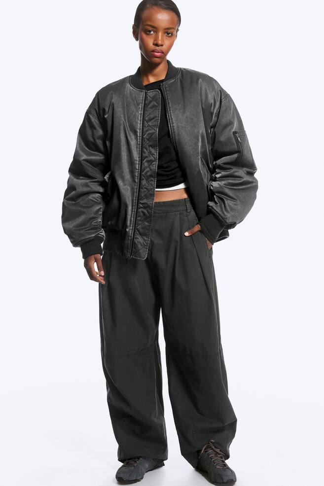 An oversized dark grey bomber jacket is worn open by the model, showcasing its ribbed collar, cuffs and hem, as well as a zipped sleeve pocket and a quilted inner placket. The jacket is layered over a black crop top and paired with wide-leg dark grey trousers. - 1