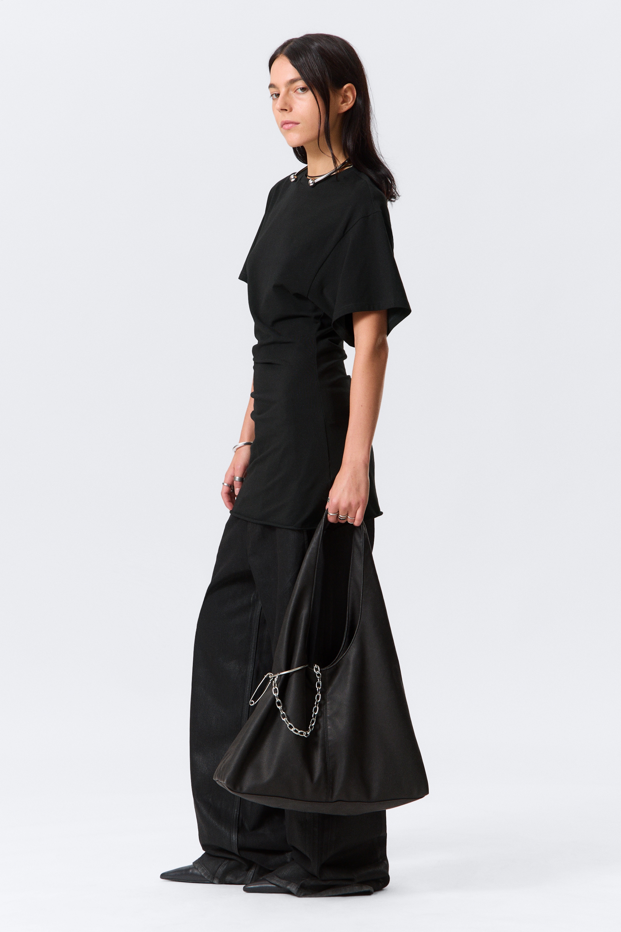 View larger image: A black hobo bag is being held, showcasing its soft, unstructured silhouette. Its integrated strap is adorned with a large silver-toned safety pin and a matching chain, highlighting its design and drape.