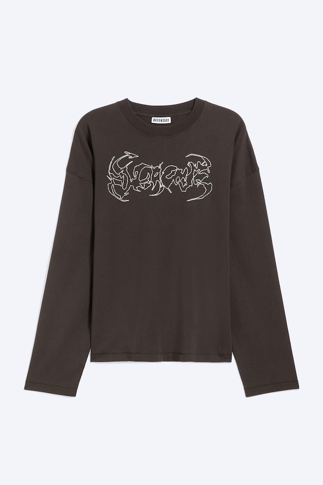 Ce t-shirt oversize à manches longues, de couleur marron foncé, est présenté de face, avec un col rond, des épaules tombantes et des manches larges à coupe droite. Un motif graphique abstrait blanc, dans une typographie dentelée de style métal, orne le centre de la poitrine. - 1