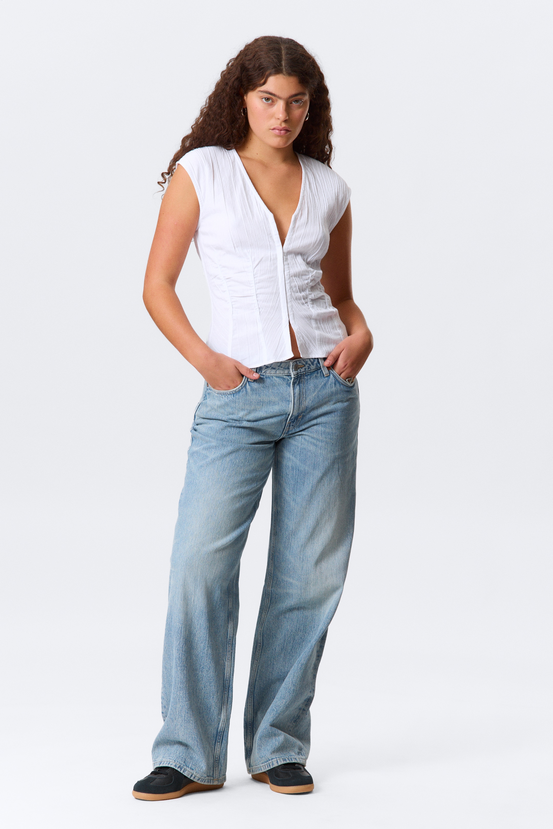 Visualizza immagine più grande: Un top bianco con scollo a V viene presentato frontalmente, caratterizzato da maniche a cappuccio, un corpetto aderente con arricciatura centrale e una texture di tessuto stropicciata. Jeans a vestibilità ampia e lavaggio chiaro completano l'outfit.