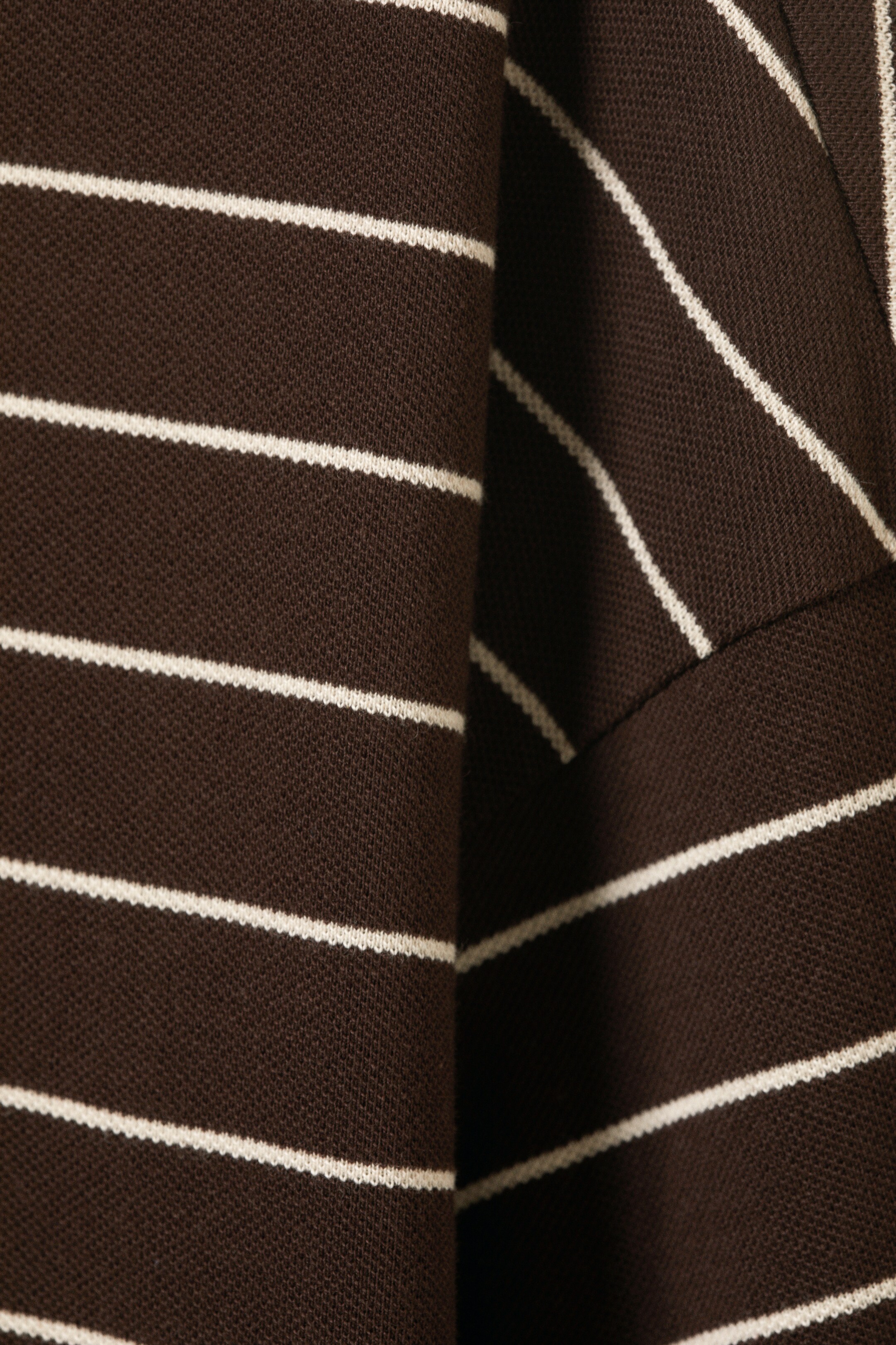 Agrandir l'image: Un polo à manches longues est présenté en gros plan, montrant son tissu marron foncé avec de fines rayures blanches horizontales. Le tissu présente une texture en maille piquée et est plié, créant une profondeur visible.